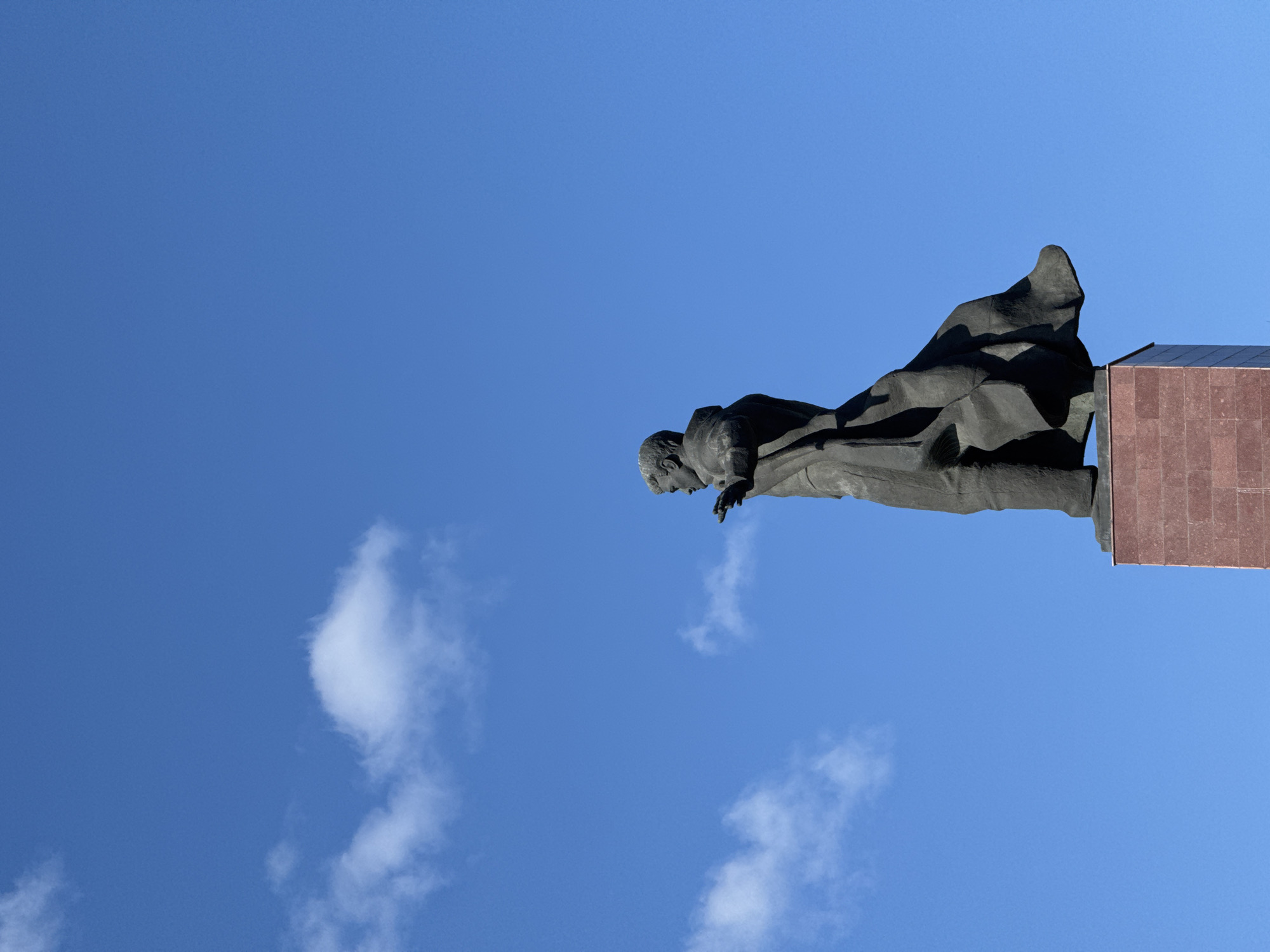A low-angle shot shows a bronze statue of Musa Cälil against a blue sky with scattered clouds. The statue is set on a red stone plinth. The photograph was taken in Kazan, Russia.