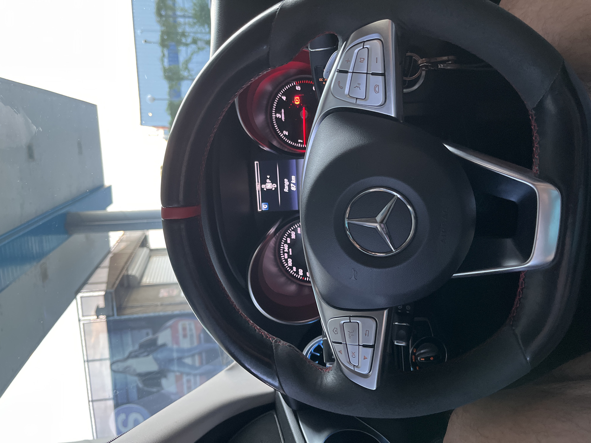 The image captures the interior of a Mercedes-Benz car from the driver's perspective, focusing on the steering wheel and dashboard. The car appears to be stationary, with the gear indicator showing "NP" (Neutral/Park) and the engine RPM needle resting at zero within the 0-5 x1000/min scale. The digital display on the instrument cluster shows a "Range" of "87 km". A red warning light, possibly indicating the parking brake or another system, is illuminated on the dashboard.
The multi-function steering wheel features a prominent Mercedes-Benz logo in the center and is adorned with red stitching. Silver-colored controls are present on both sides of the steering wheel. The right side includes buttons for volume (+/-), phone (answer/hang up), and menu navigation, while the left side has directional arrows, "OK", "OFF", and a home button. Text "AIRBAG" is visible on the steering wheel. Car keys are hanging from the ignition or a nearby hook.
Through the windshield, bright daylight indicates it is daytime. Reflections of an outdoor environment are visible, including what appears to be parts of buildings or structures, possibly a canopy or wall, with indistinct signs showing letters such as "S" and "A". The speedometer scale is visible on the right, showing numbers up to "200" km/h. The overall scene suggests the car is parked outdoors, potentially near a commercial area in Shumen, Bulgaria.