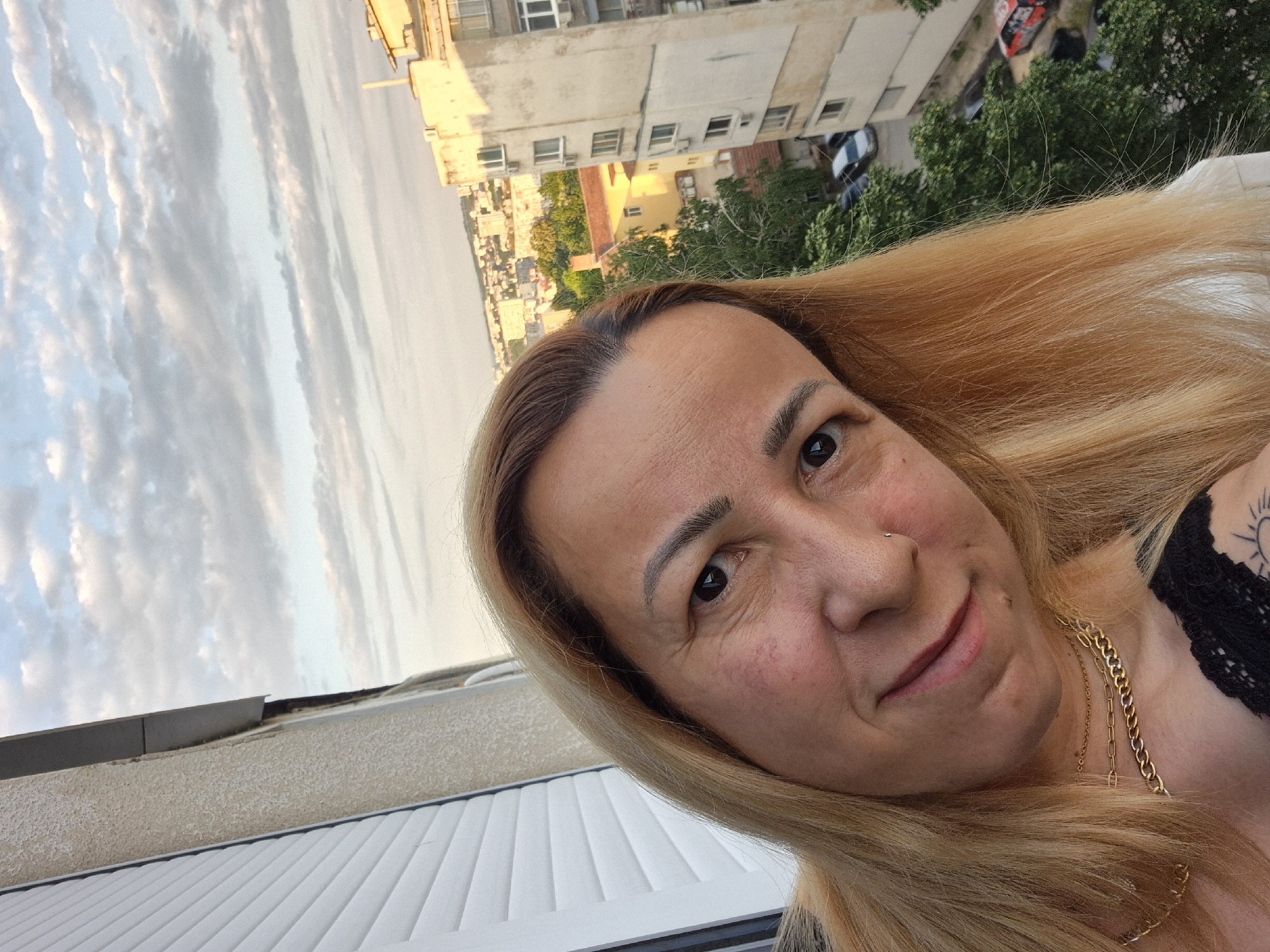 A woman is taking a selfie on a balcony with a city view. She has blonde hair and is wearing a gold necklace and a black lace top. The background shows buildings and trees under a cloudy sky.