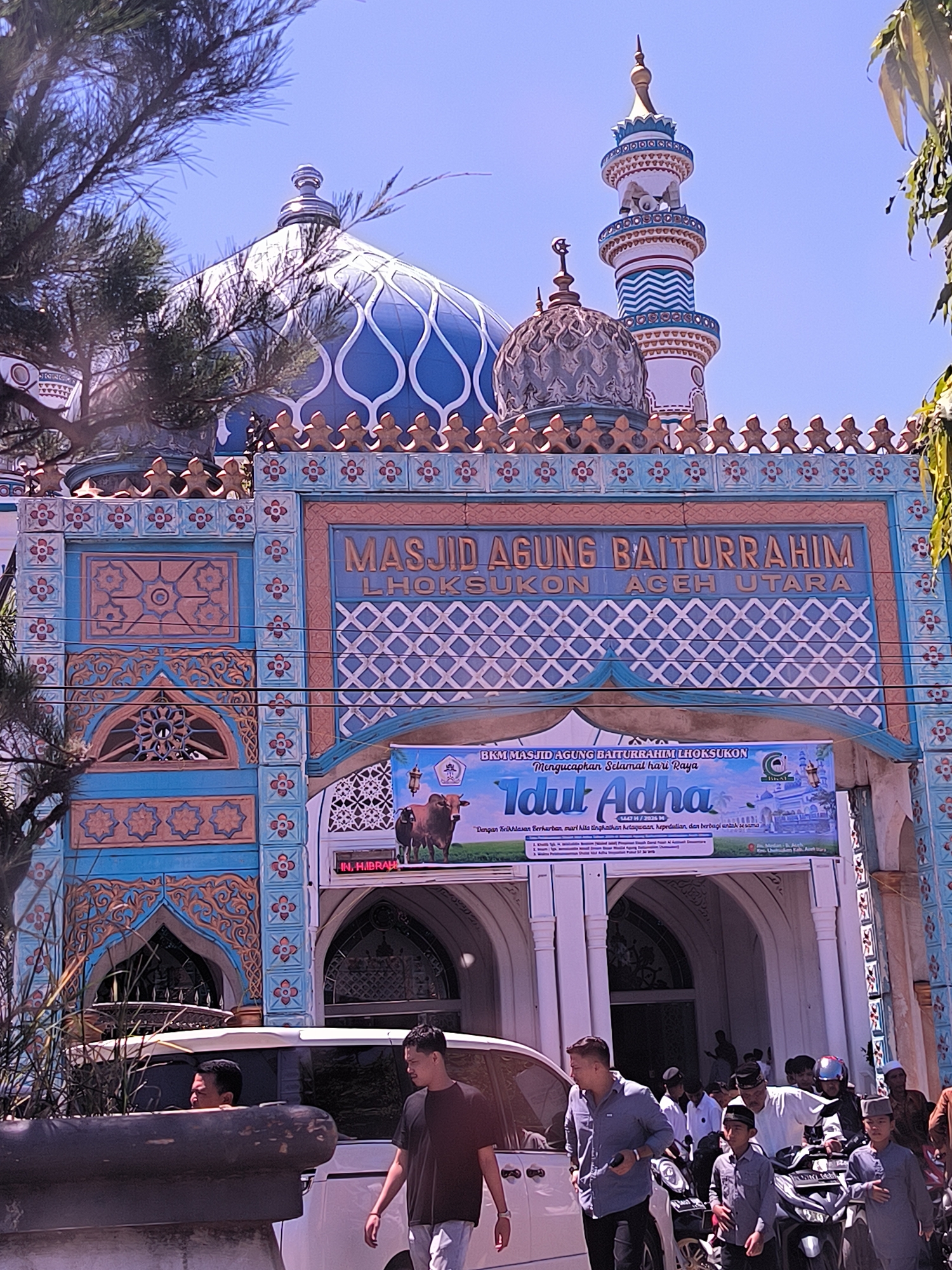 Here is a description of the image:
The image shows the Masjid Agung Baiturrahim mosque in Lhoksukon, Aceh Utara, Indonesia. The mosque's facade is adorned with blue and orange geometric and floral patterns. A large blue dome with white accents sits atop the mosque, with a minaret rising behind it. A banner promoting "Idul Adha" hangs above the entrance.
In front of the mosque, a group of people are gathered, including men, women, and children. Several motorcycles are parked nearby, and a white van is partially visible on the left. The foreground is slightly obstructed by tree branches. The weather appears sunny.