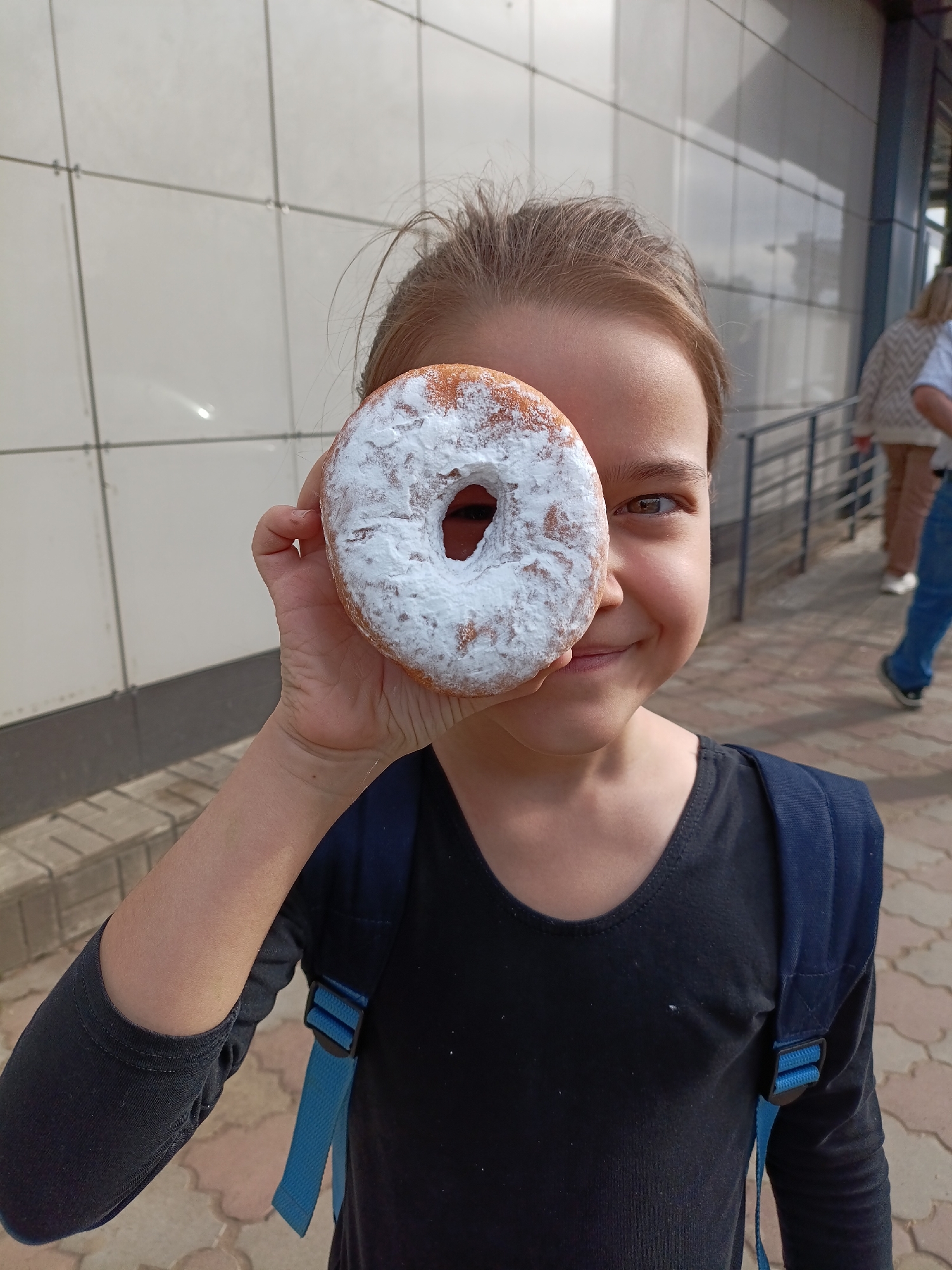 A young girl holds a powdered donut up to her eye, obscuring it. She is smiling. She is wearing a dark long-sleeved shirt and a blue backpack. The background shows a building with a white tiled facade and a metal railing. Other people are visible walking in the background. The location is outdoors, near a building with sidewalk bricks visible. The city is Chaikovsky, Russia.
