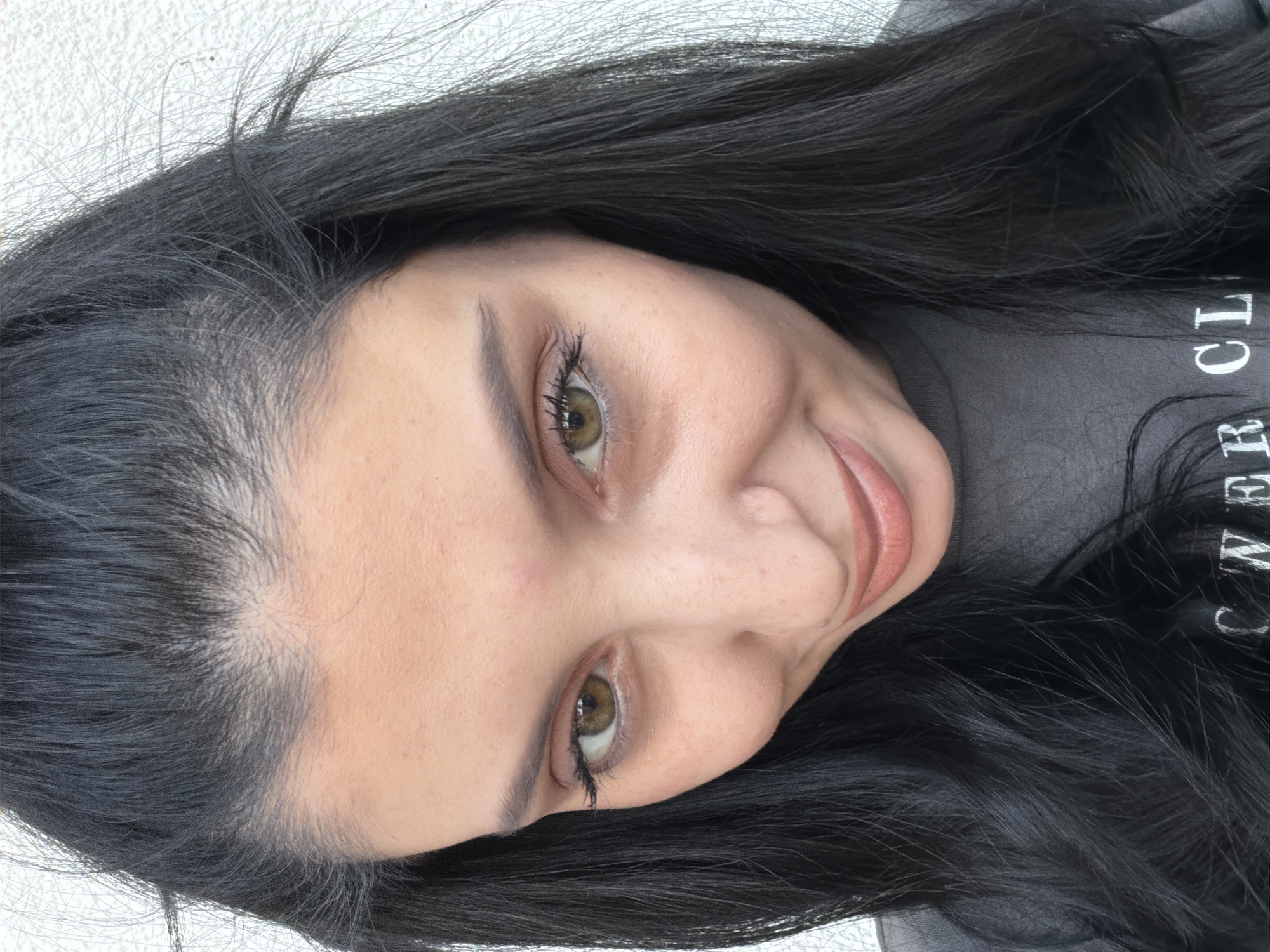A close-up, head-and-shoulders portrait shows a woman with dark hair and light skin against a white wall. She is looking down and to her left with a slight smile. The woman has brown eyes, dark eyebrows, and light pink lipstick. Her dark hair is parted in the middle and falls around her face and shoulders. She is wearing a grey t-shirt. The background appears to be a textured white wall, suggesting an outdoor setting in a city. The image quality is clear, with natural lighting. There is no identifiable information suggesting the location is Tbilisi, Georgia.