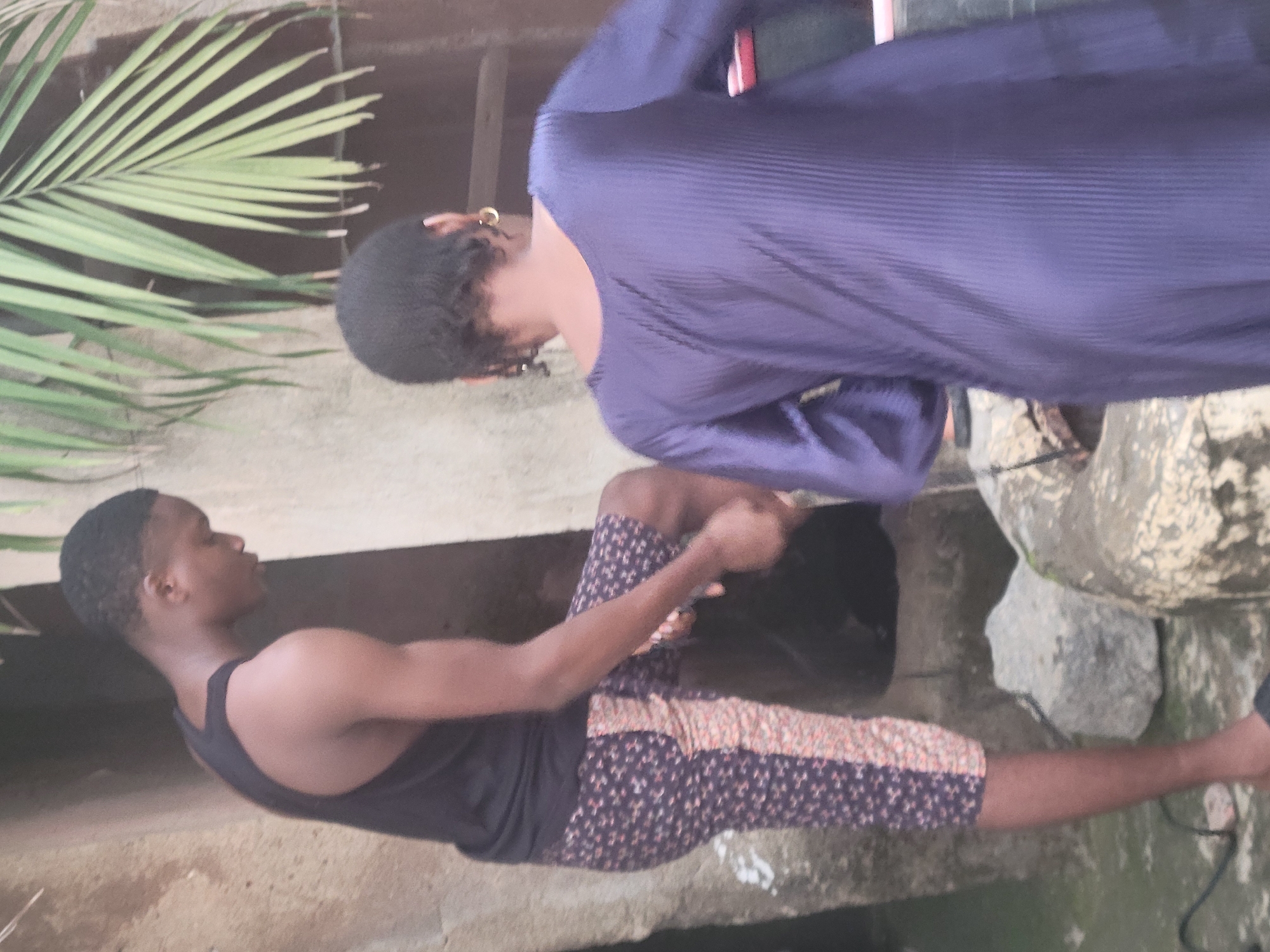 In Madalla, Nigeria, two individuals are captured in a candid moment. On the left, a young man, dressed in a black tank top and patterned shorts, is looking down. His right leg is bent and raised, as if he is in the process of stepping over something. The shorts have a dark blue base with a small, repeating floral pattern in white and pink, with a vertical stripe of a lighter, peachy-orange fabric on the outer side of his right leg.
To the right, a person from behind, wearing a long-sleeved, dark purple dress with a ribbed texture, is facing away from the camera. They are positioned slightly behind and to the right of the man. Their hair is short, and they are wearing a hoop earring on their visible ear. The background suggests an outdoor setting, with textured walls and a large palm frond visible at the top of the frame. Rocks and what appears to be a rudimentary structure or opening are also part of the environment. A black cable is visible on the ground near the man's feet. The lighting indicates it is daytime, with natural light illuminating the scene.