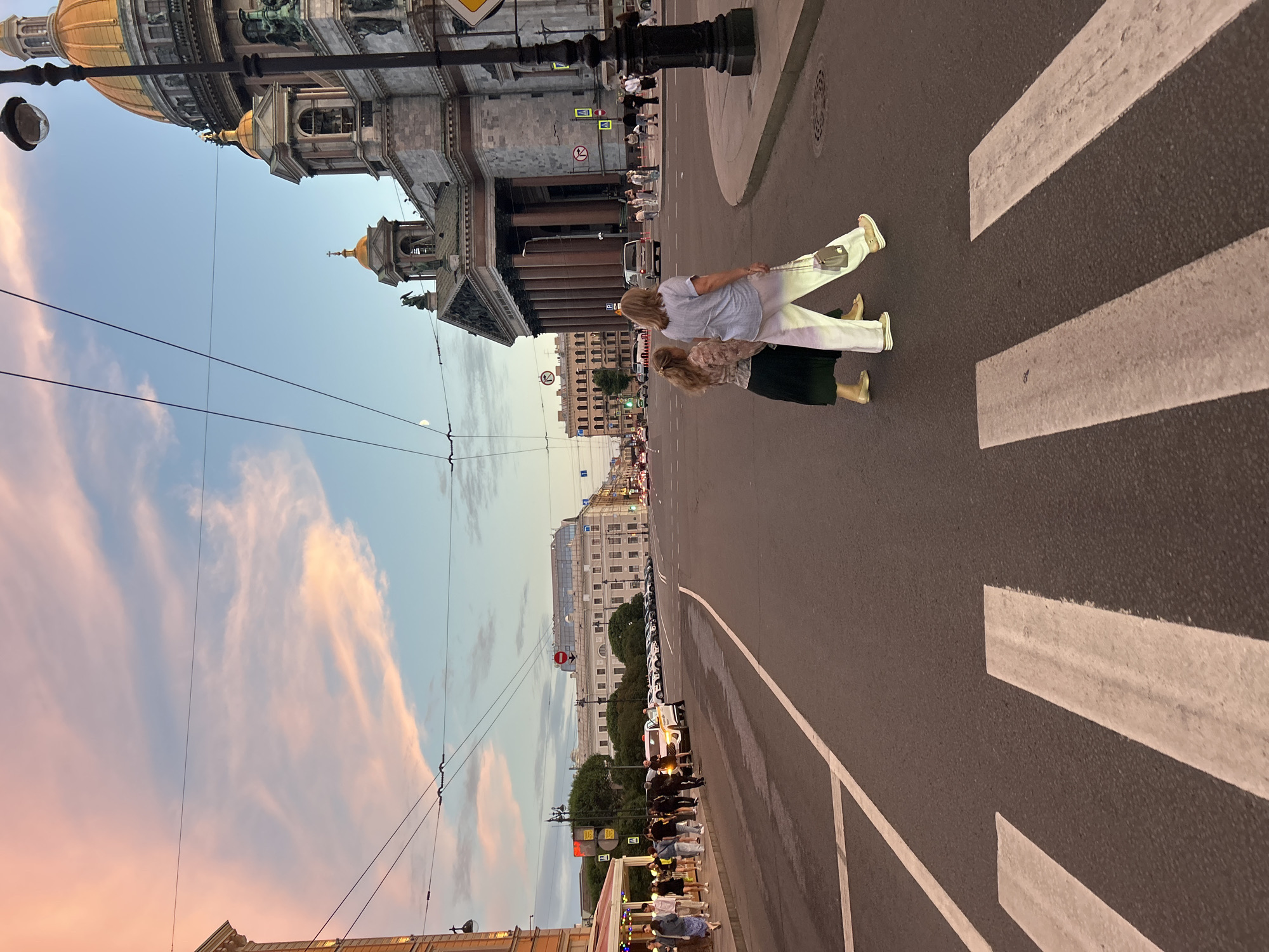 The image depicts a street scene at dusk in a European city, likely St. Petersburg, Russia, with St. Isaac's Cathedral prominently featured on the right. Two women are crossing a crosswalk in the foreground, walking from right to left. The woman on the left wears a dark skirt and a patterned blouse, while the woman on the right is dressed in white pants and a light blue top, carrying a small gray purse.
The street is wide, with multiple lanes of traffic. Cars are parked along the side of the road in the distance, and a white taxi is visible. A line of pedestrians walks on the sidewalk to the left, some of them are entering or exiting a building with outdoor seating. Overhead, power lines crisscross the sky.
The sky is a soft blend of pink, orange, and blue, indicating the sunset. The architecture in the background is grand and classical, characteristic of historic European cities, with a large, ornate building and a domed cathedral. Streetlights are present, with one in the foreground casting a light. The overall atmosphere is calm and serene, with the soft lighting and the muted colors of the sky.