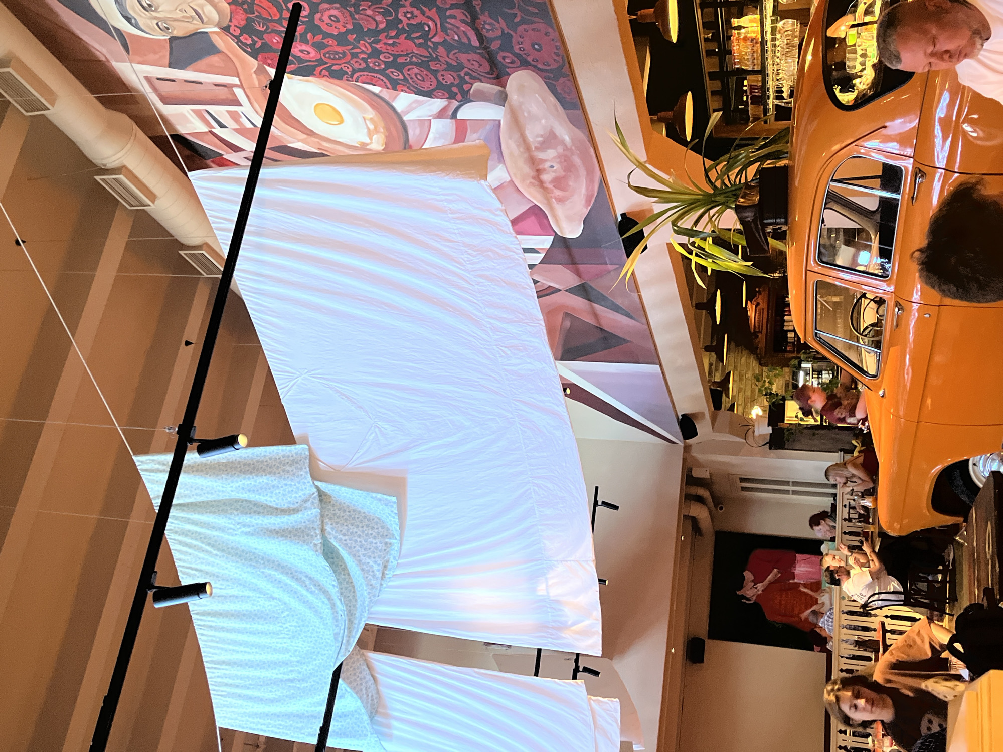 The image shows the interior of a restaurant. In the foreground, an orange vintage car is parked, with a plant on its roof. The car is a focal point of the scene. The background features a large mural on the wall depicting two figures in red attire. Hanging from the ceiling are several large white fabric sheets, creating a draped effect. The restaurant appears to be busy, with several patrons seated at tables. Lighting fixtures are visible on the ceiling and walls. The overall ambiance suggests a trendy and eclectic dining establishment.