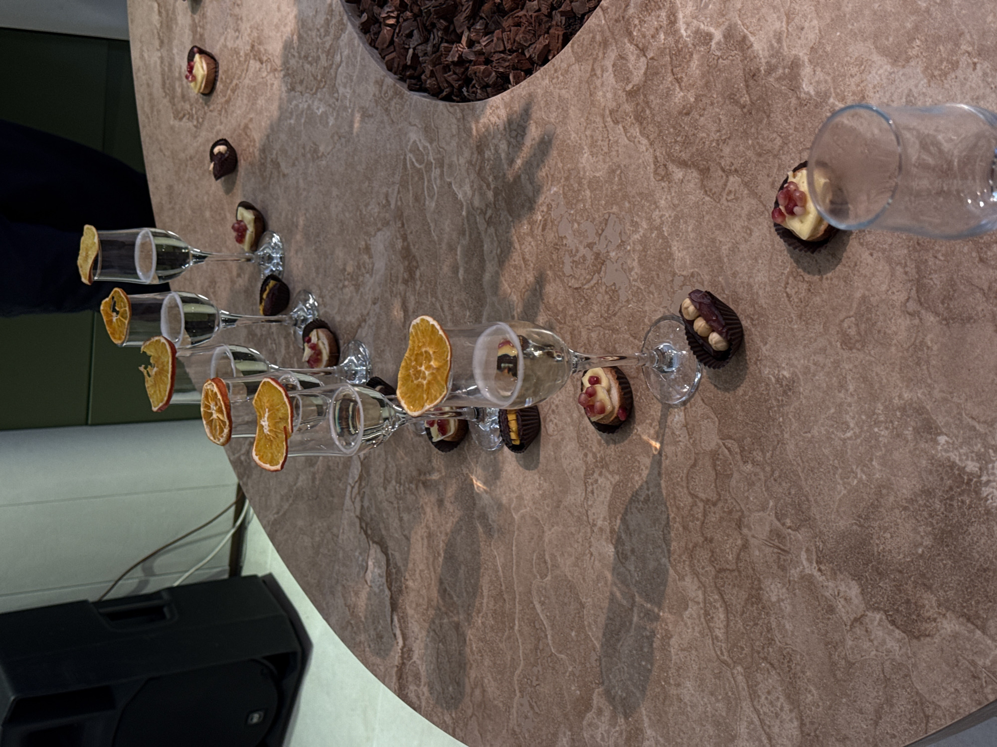 The image captures a close-up, high-angle view of a contemporary event setup, likely in Yerevan, Armenia. The central subject is a large, irregularly shaped table with a textured, brownish-grey stone or tiled surface. A curved cut-out in the table's center is filled with dark, bark-like material, possibly decorative.
Arranged in a line across the table are several champagne flutes, each holding a dried orange slice as garnish. Most flutes appear to contain a beverage, possibly champagne. Accompanying the glasses are small, individual desserts or tarts in dark paper wrappers. These desserts vary, some featuring a light-colored cream filling topped with red fruit (possibly pomegranate seeds), while others are dark chocolate confections. One empty, clear drinking glass lies on its side towards the top right.
A person wearing dark clothing is partially visible on the far left edge of the image, suggesting human presence at the event. In the bottom left corner, part of a black speaker and some cables are seen, indicating audio equipment for an event. The overall scene suggests a sophisticated gathering, such as a reception, tasting, or a celebratory event. The indoor setting features muted tones, with a dark green or blue wall in the background. No specific time of day or weather conditions are discernible, and no visible text is present.