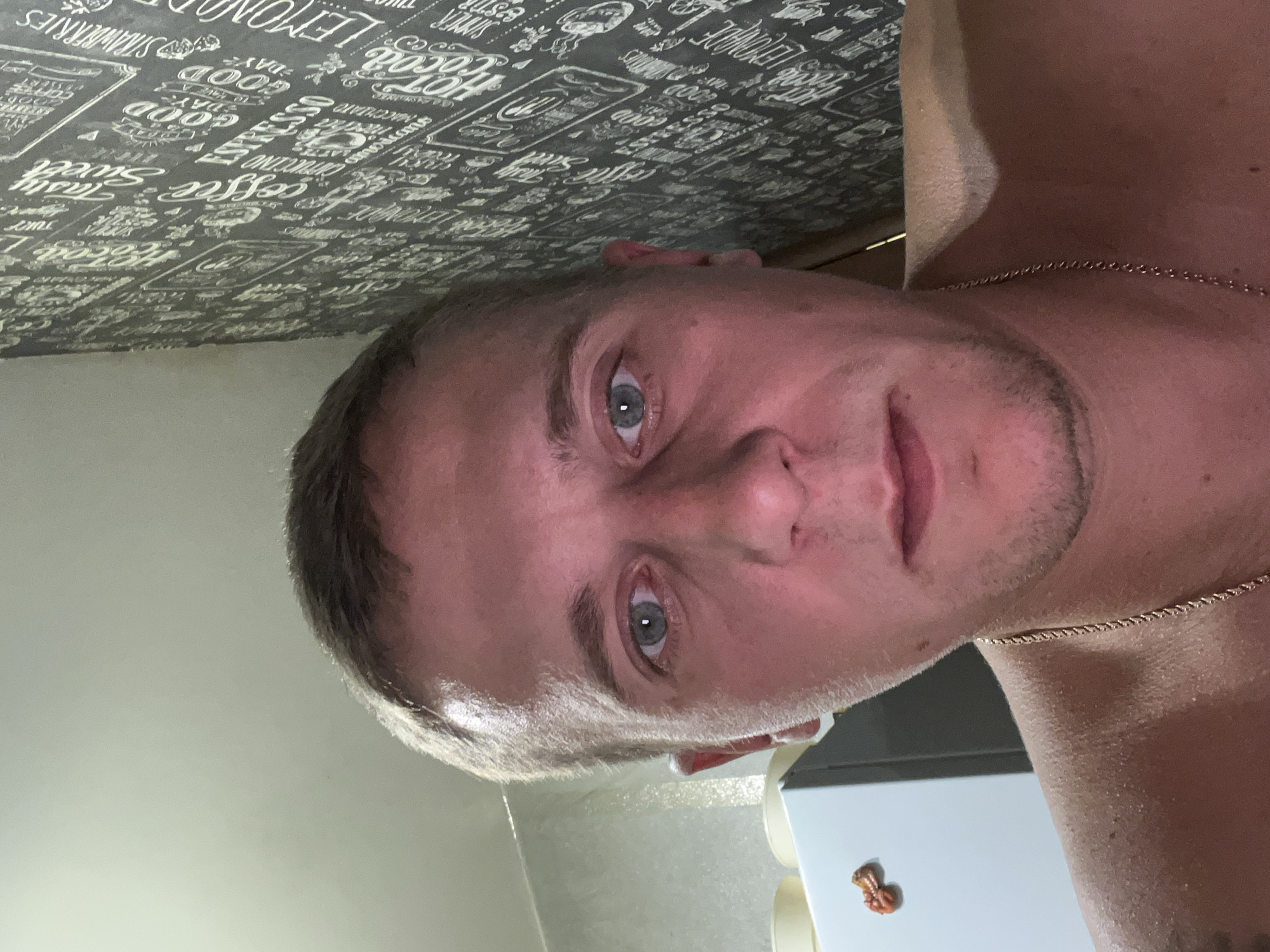 A close-up, selfie-style portrait captures a Caucasian male in his late 20s or early 30s, with light skin and short, light brown hair. He is facing the camera directly, with his shoulders visible. The man appears to be indoors, with a textured, dark wallpaper featuring white text and graphics on the right side of the frame. To the left, a portion of a white refrigerator is visible, with a small, red octopus-shaped magnet attached to it. The lighting in the scene is warm and appears to come from the front, casting a pinkish hue on the man's skin. The man is wearing a gold chain around his neck. The background wallpaper has text that appears to be related to food and beverages, with words like "Coffee," "Espresso," "Lemonade," and "Strawberries" discernible. The image does not provide any specific indicators of its location within the city of Minsk, Belarus.