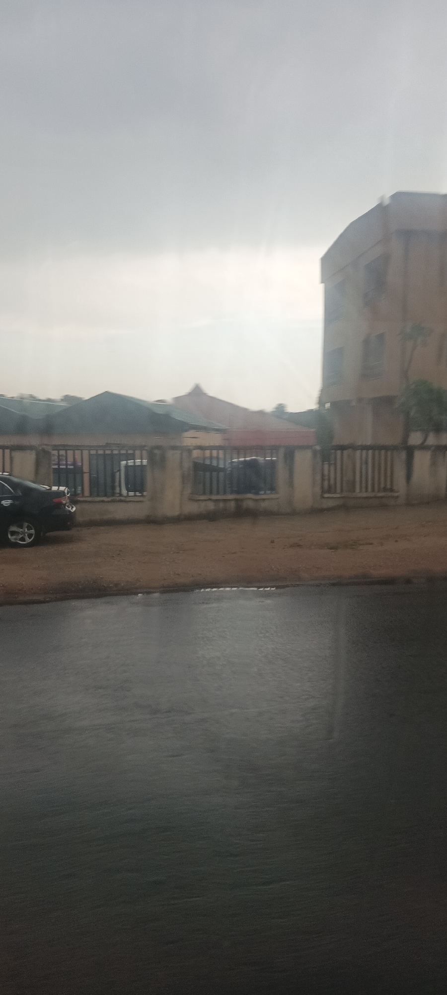 The image shows a street scene in Jos, Nigeria on a cloudy day. The road in the foreground is wet, indicating recent rain. A dark-colored car is partially visible on the left, parked behind a low wall with vertical bars. Behind the wall, there are other vehicles parked. In the background are single-story buildings and a taller, tan-colored building with multiple stories on the right. The sky is overcast and grey.
