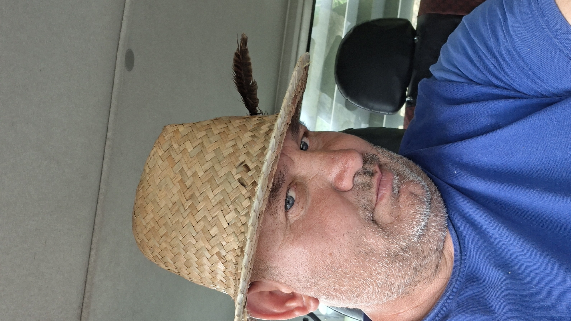 A close-up, selfie-style photograph shows a man wearing a straw hat with a feather in it, and a blue t-shirt. He has light blue eyes, grey stubble, and is looking directly at the camera. The background appears to be the interior of a vehicle, with a headrest of a black seat visible behind him. The man's left ear is clearly visible above the brim of the hat. The image was taken in the city of Hîncești, Moldova.