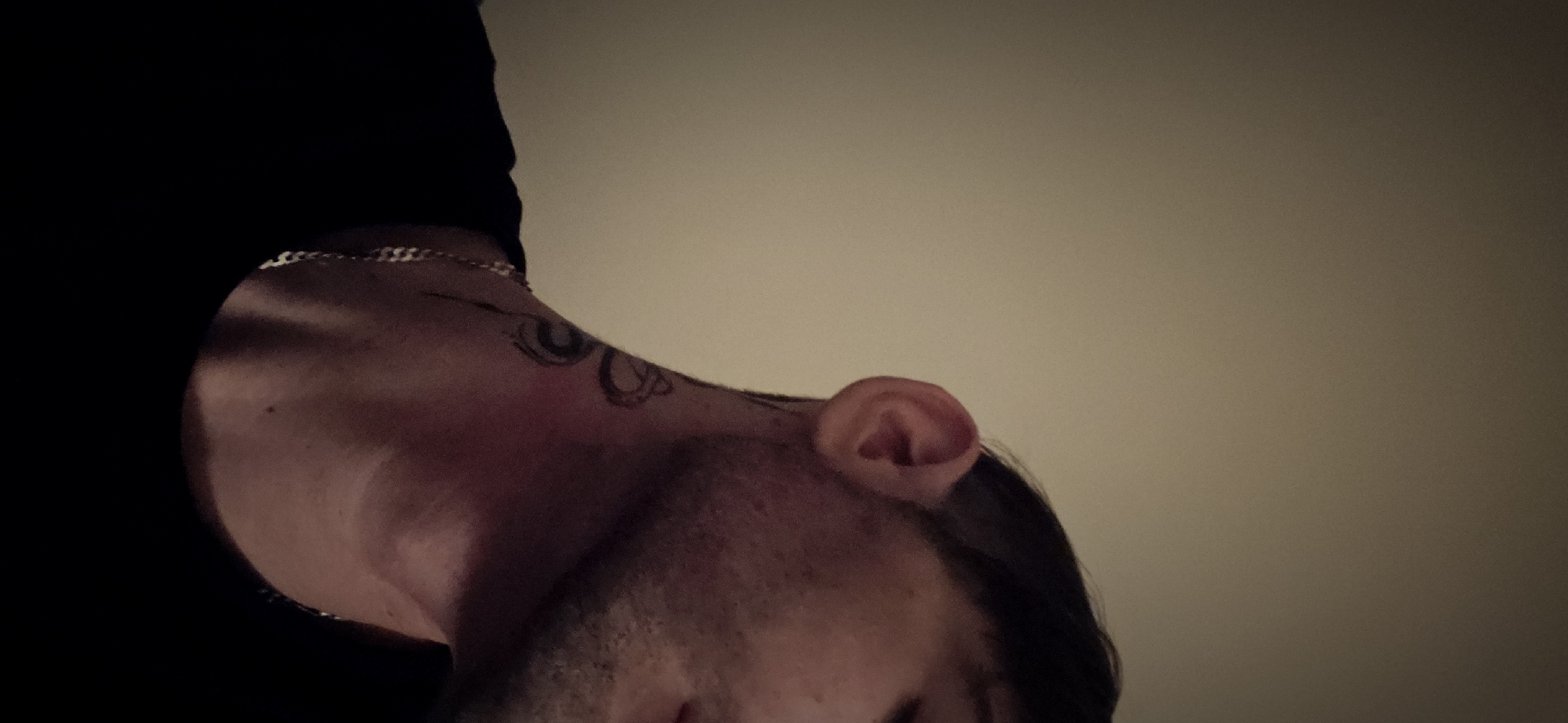 The image presents a close-up, low-angle shot of a person's head and neck, looking downwards. The subject appears to be male, with dark hair and some facial hair visible. They are wearing a dark top, likely a t-shirt, and a silver-colored chain necklace is visible around their neck and shoulder.
A notable feature is a dark-inked tattoo on the left side of the person's neck, featuring an intricate, possibly abstract design that seems to incorporate circular or eye-like elements. The person's left ear is also visible in the upper right portion of the frame.
The background is a plain, light, solid color, resembling an indoor wall or ceiling. The lighting is soft and appears somewhat dim, suggesting an indoor setting potentially during evening hours or in a subdued environment. The photo's perspective is from below, looking up at the subject, creating an intimate and somewhat abstract view. No specific activity beyond the downward gaze is discernible, and there is no visible text.