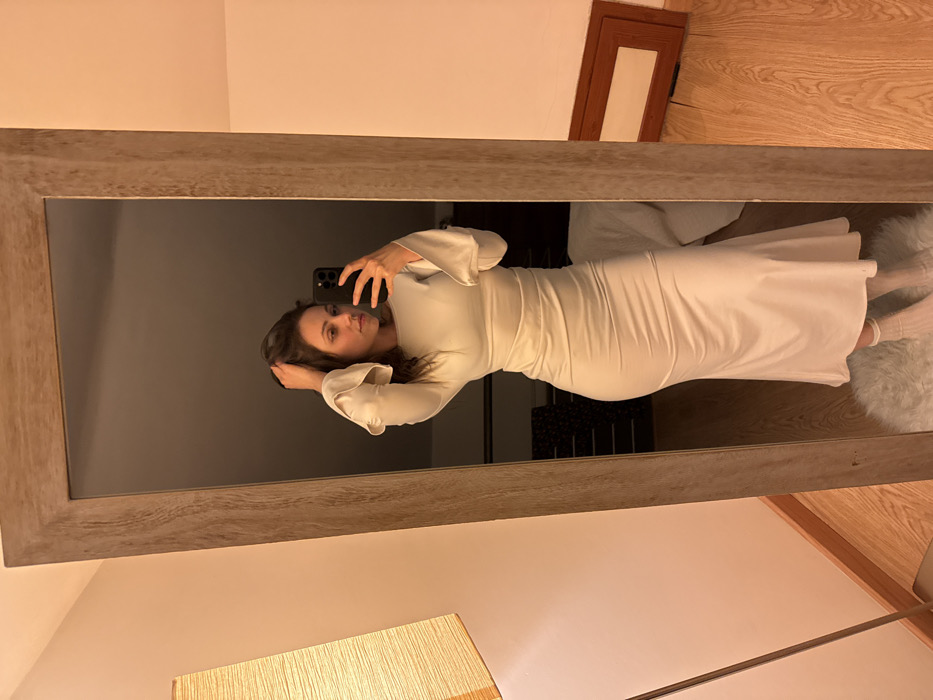 The image displays an indoor scene, likely a residential bedroom or dressing area, featuring a female individual captured in a full-length mirror selfie. The specific location within Cava de' Tirreni, Italy, cannot be determined from the visual evidence presented in the image.
The subject, an adult female with dark hair, stands facing the mirror, holding a dark-colored smartphone with a visible multi-lens camera array in her right hand to record the image. Her left hand is positioned in her hair near her head. She is wearing a light-colored, long-sleeved midi-dress that is form-fitting through the bodice and hips, with a boat neck, ruched detailing around the waist, and flared cuffs. Light-colored socks or foot coverings are visible on her feet.
The mirror itself features a wide, light-toned wooden frame with visible grain. To the left of the mirror, a floor lamp with a tall, cylindrical beige fabric shade is present. A white, shaggy rug occupies the floor directly beneath the individual, while the visible flooring outside the rug and in the background reflection is light-colored hardwood. The walls in the reflection are a light, solid color. In the reflection to the right of the individual's lower body, a brown rectangular frame or object is leaning against a wall. The overall lighting is warm, suggesting artificial light sources.
