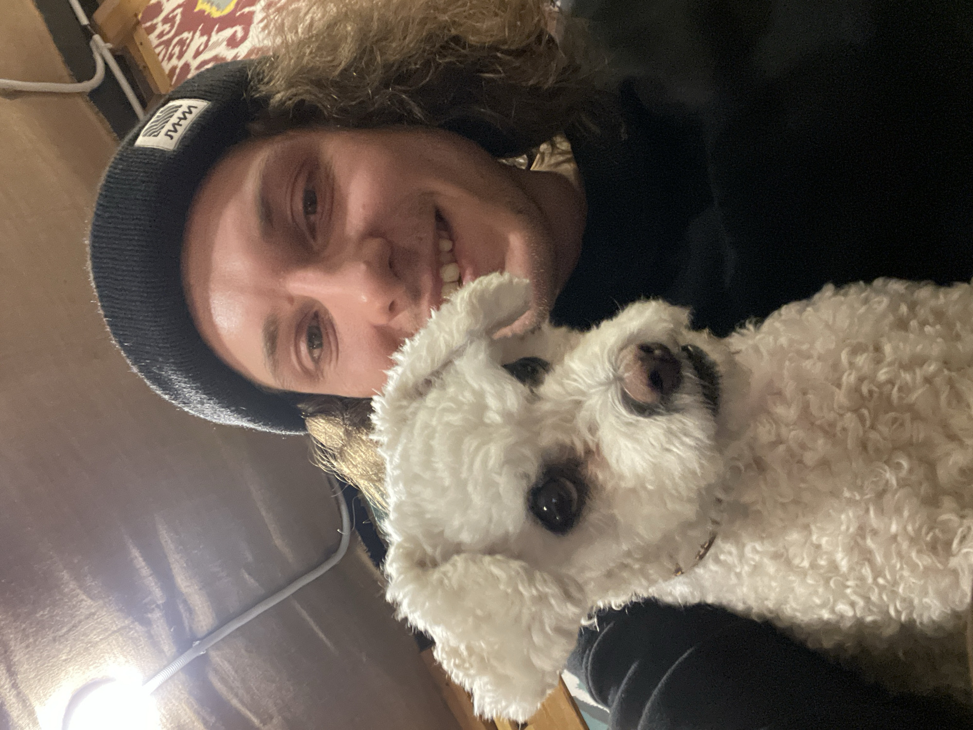 A selfie photograph shows a man with curly blond hair wearing a black beanie with a white logo, holding a small white dog. The man is smiling, and the dog is looking at the camera. The background is indistinct but appears to be an interior space with a wooden wall and a patterned fabric visible in the upper left corner. There is no visual information suggesting the location is Yekaterinburg, Russia.
