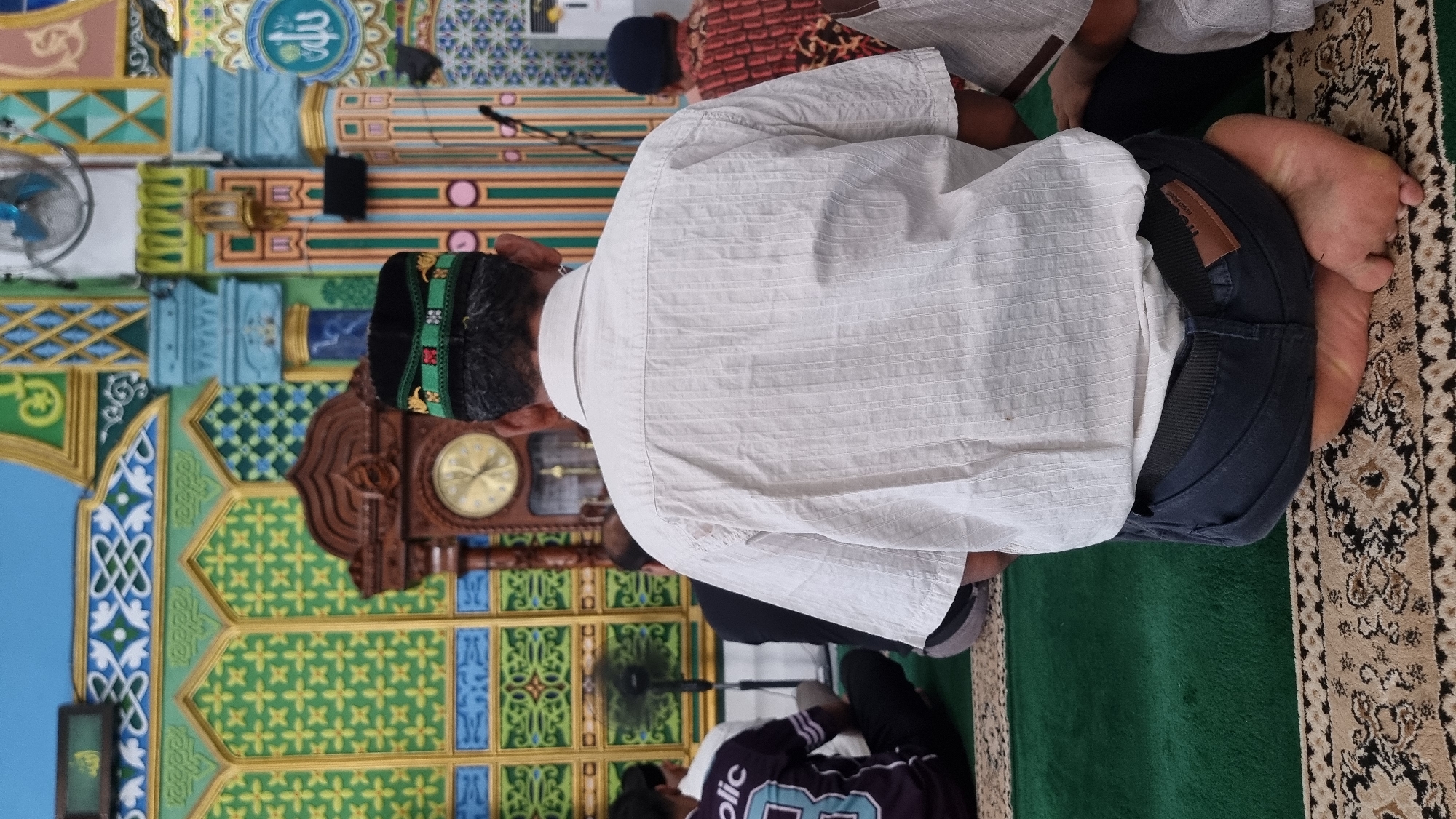 In the city of Keudekruenggeukueh, Indonesia, a man wearing a white short-sleeved shirt and dark pants is kneeling on a patterned rug inside a mosque. The man is facing away from the camera, and his head is covered with a black kufi hat adorned with green and gold embroidery. Behind him, another man in a red and black patterned shirt is also kneeling. The mosque's interior features vibrant green and yellow walls decorated with intricate Islamic geometric patterns. A large, ornate wooden clock with a gold face is mounted on the wall. Several fans are visible, one of which is actively rotating, suggesting a warm climate. The lighting appears to be artificial.