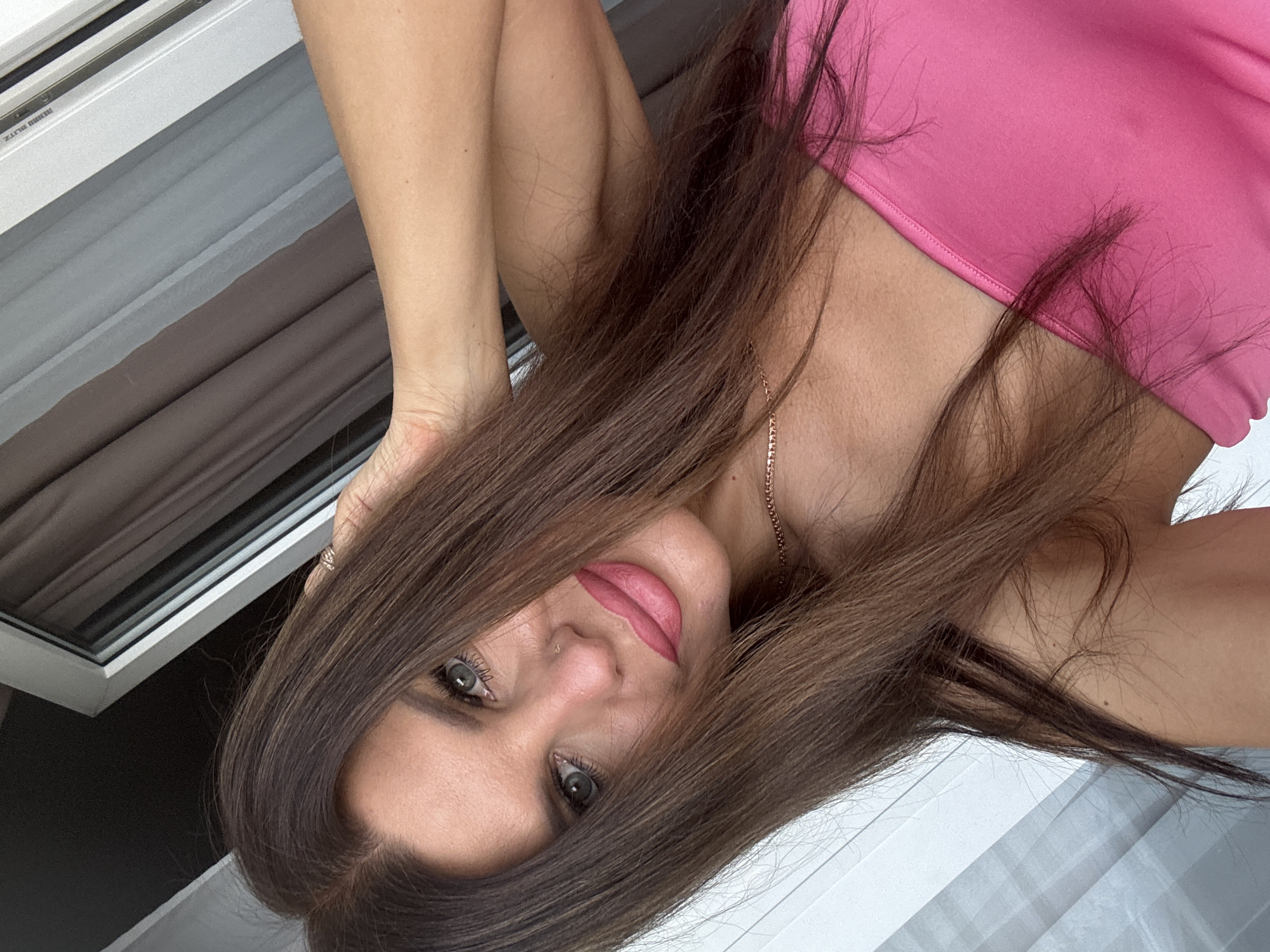 A close-up, angled selfie of a woman in a bright pink strapless top. Her long, dark brown hair is partially obscuring her face and is being held back by her left hand, which is positioned behind her head. She has blue-grey eyes, a small nose piercing, and her lips are colored with a matte pink lipstick. A delicate gold necklace is visible around her neck. The background consists of a window with sheer and opaque curtains, suggesting an indoor setting, possibly a hotel room or apartment. The lighting is natural and appears to be coming from the window. The scene does not contain any recognizable landmarks or features that would indicate a specific location within the city of Chaikovsky, Russia.