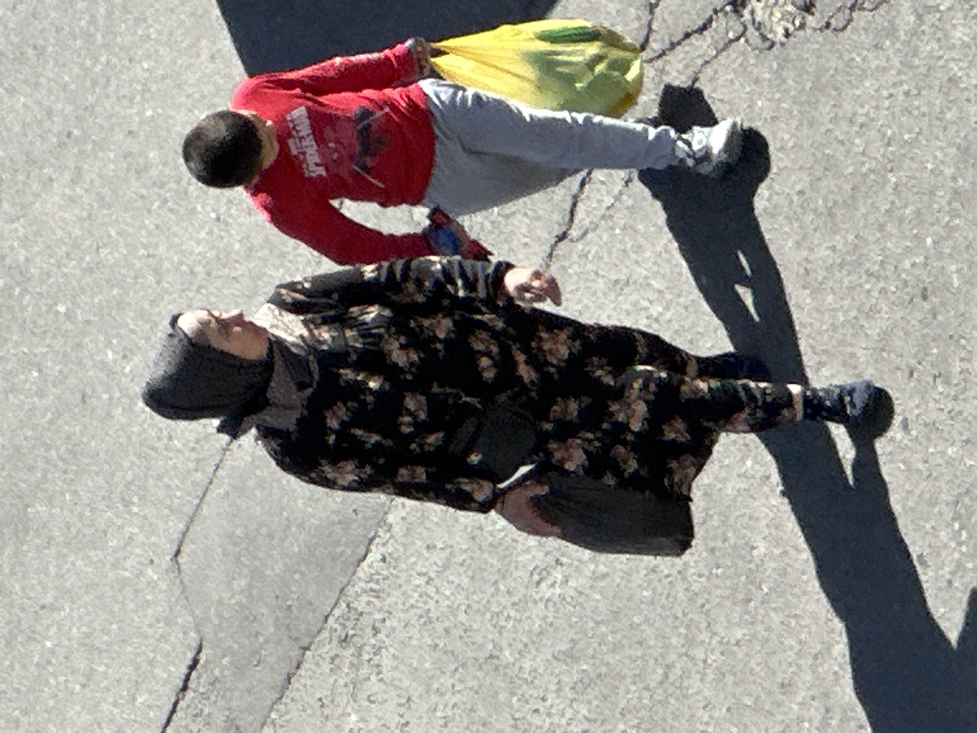 Two individuals, an adult woman and a young boy, are walking on a paved surface, possibly a sidewalk or street, in Podolsk, Russia. The woman is on the left, wearing a dark floral patterned dress and a headscarf. She is carrying a dark bag in her left hand. The boy is on the right, wearing a red long-sleeved shirt with a Spider-Man graphic and light gray pants. He is carrying a bright yellow shopping bag filled with produce in his right hand. The ground is concrete with visible cracks and patches of dirt. Sunlight casts distinct shadows of the individuals on the ground.