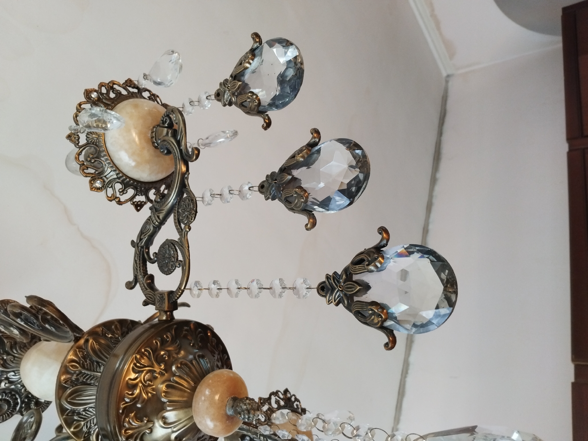 This is a close-up, eye-level shot of a decorative chandelier made of antique brass and clear crystal teardrop pendants. The chandelier is suspended from a white ceiling. The focus is on the ornate metalwork, which features intricate floral and leaf motifs, and the faceted crystal drops that dangle below. One of the crystal pendants is larger and more prominent than the others, positioned in the lower left of the frame. The background consists of the ceiling and a portion of a wall, showing some minor imperfections and a decorative crown molding. The image appears to have been taken indoors, likely in a residential or public space. There are no people or specific landmarks visible in the image to indicate its location in Akhaltsikhe, Georgia.
