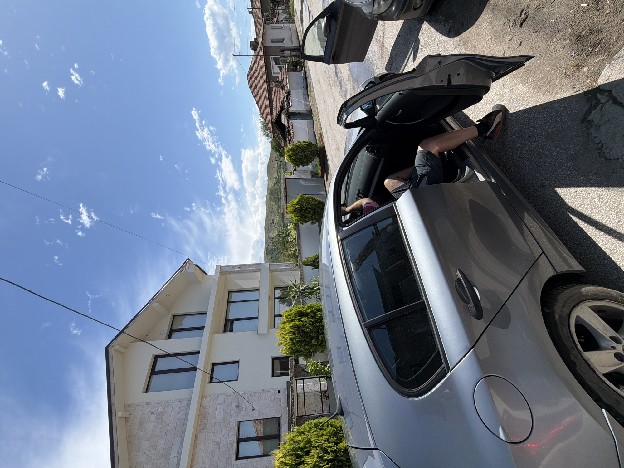 This vertically oriented image, presented as if rotated 90 degrees counter-clockwise, captures a bright, sunny daytime scene on a street, consistent with a residential area in Sinitovo, Bulgaria.
In the mid-ground and foreground, a silver-grey sedan car is parked on an asphalt road. Its front passenger door is wide open, with a person's lower legs and feet, clad in black shorts and a black shoe with red detailing, visible inside the vehicle. The driver's side door is also ajar. Strong, dark shadows on the asphalt suggest direct, intense sunlight, likely around midday or early afternoon. A second, darker-colored car is partially visible in the upper right, also seemingly parked.
To the left of the parked car, a prominent, multi-story modern building with a light-colored exterior and dark-framed windows stands. Its lower levels are faced with light-colored stone. Further down the street, behind this modern structure, an older, more traditional building with a red tile roof and white walls is visible. Several neatly trimmed green bushes or small trees are planted along the side of the modern building and between the structures.
The background reveals distant, undulating hills or mountains under a vibrant blue sky dotted with scattered white cumulus clouds. Power lines traverse the upper portion of the frame. The scene portrays a blend of architectural styles in a town setting, nestled within a hilly landscape. No discernible text is visible in the image.
