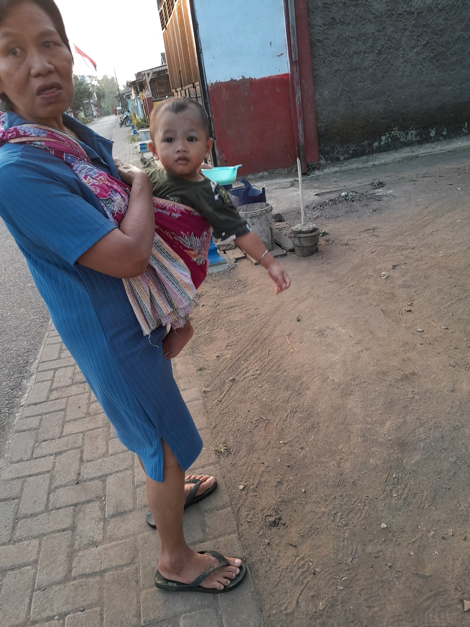 An elderly woman, wearing a blue dress and a colorful scarf, carries a young child on her back in a patterned sling. The child wears a green shirt and is visible from the waist up. They are standing on a brick sidewalk next to a dirt road. In the background, there are buildings, a street with some debris, and a flag. The scene appears to be outdoors in a semi-urban or rural area. The overall impression is of a daily activity, likely in the city of Ngadilangkung, Indonesia, given the context.