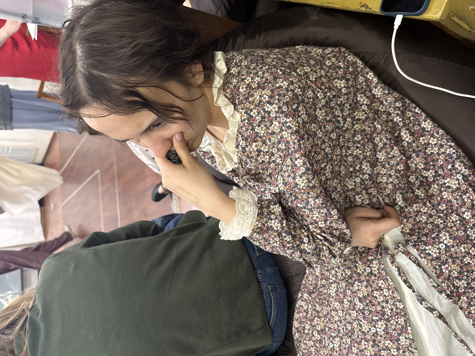 A woman with dark hair and pale skin is seated, wearing a long-sleeved floral dress with a lace-trimmed collar and cuffs. She's holding a small, dark object near her face, possibly examining it. To her left, another person is partially visible, wearing an olive green shirt and blue jeans. The floor is tiled in reddish-brown. A phone is plugged into a charger on a surface to the right.
I am unable to make a judgement about the specific location as I lack any additional information about the scene.