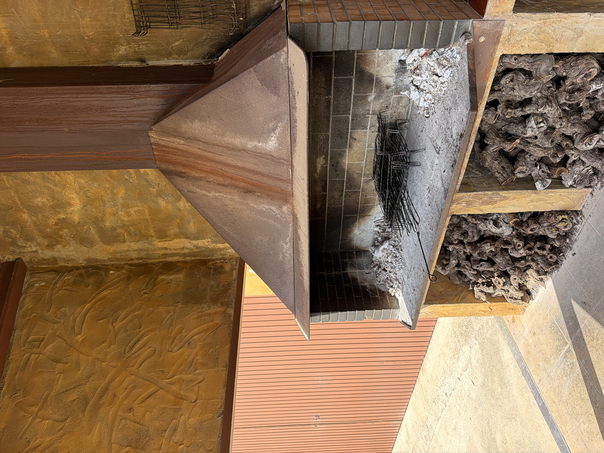 This image captures an outdoor barbecue or fireplace setup, likely on a patio or terrace, in Bonastre, Spain. The central feature is a robust structure with a large, weathered, rusty metal hood and chimney. Below the hood is a brick-lined firebox, filled with a considerable amount of white ashes and charcoal debris, along with a metal grate, suggesting recent use.
To the right of the firebox, a built-in cubby or storage area holds a large stack of dark, gnarled wood pieces, possibly grapevines, ready for fuel. The surrounding architectural elements include a textured, ochre-colored stucco wall on the left, a dark brown wooden beam running horizontally across the top, and a reddish-brown slatted surface in the foreground, which could be decking or a path. Light-colored tiled or concrete flooring extends to the right of the barbecue area.
The scene is brightly illuminated by natural daylight, casting distinct shadows and indicating a clear, sunny day. No people are present, and no activity is currently taking place. There is no visible text in the image. The overall aesthetic is rustic and functional.