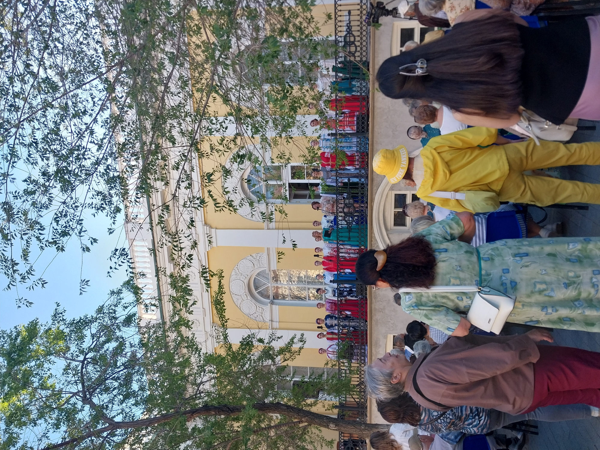 The image shows a group of people standing on a balcony of a yellow building in what appears to be Orenburg, Russia. The building has ornate white detailing around arched windows. The people on the balcony are wearing traditional Russian clothing in a variety of colors. In front of the building, there are trees and people standing in the street looking up at the balcony. Some of the people in the crowd are wearing casual clothing. The sky is blue.