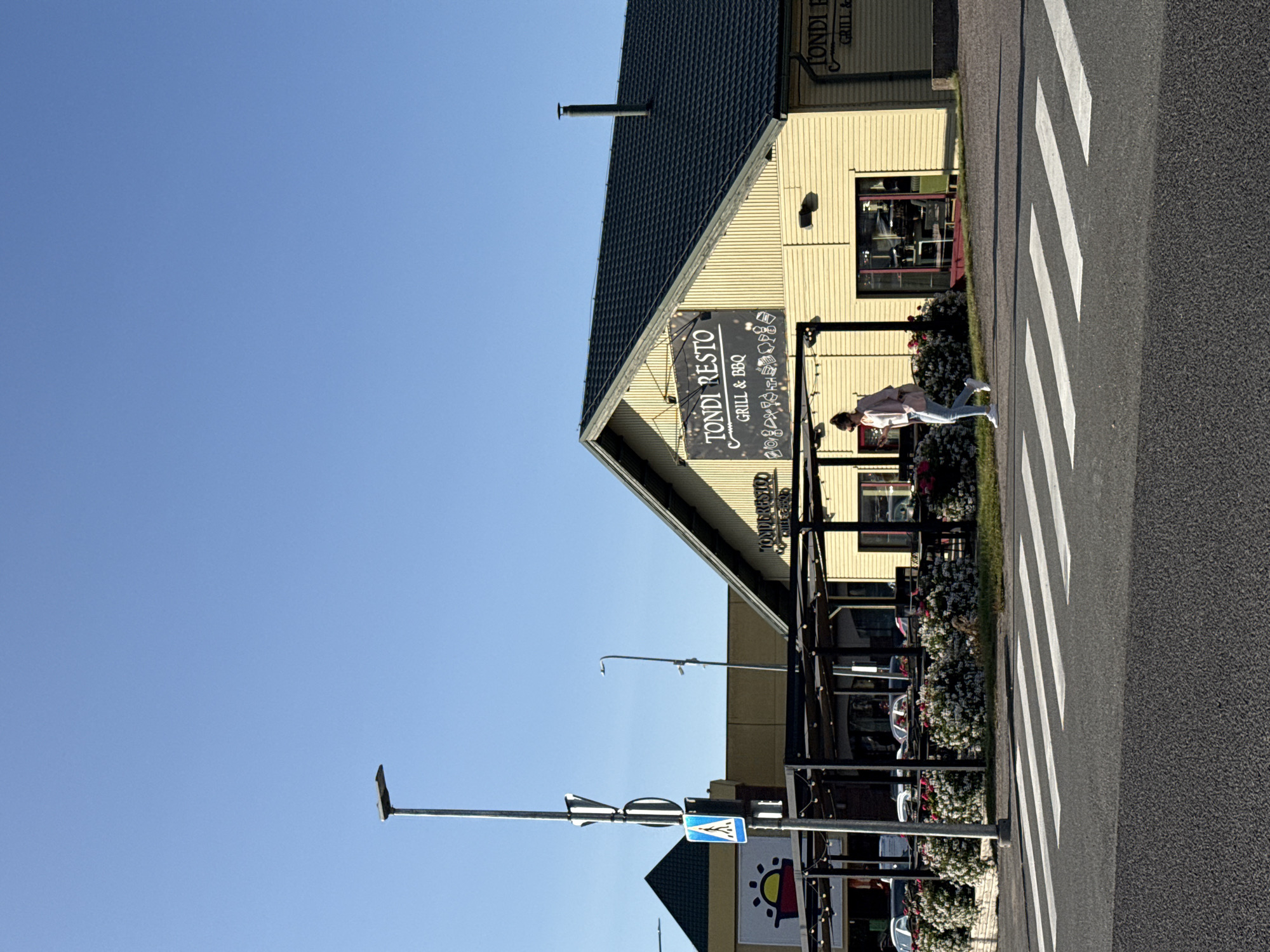 A woman walks across a crosswalk in front of a yellow building with a dark roof. The building has a sign that reads "TONDI RESTO GRILL & BBQ". The building is likely a restaurant. There are tables and chairs set up outside under a dark awning with string lights. There are flower bushes in front of the awning. A street light is visible to the left of the building. The sky is clear and blue. The image appears to be taken during the daytime. The scene is likely in a commercial or suburban area. Based on the text on the sign, the location is likely Tondi Resto in Tallinn, Estonia.