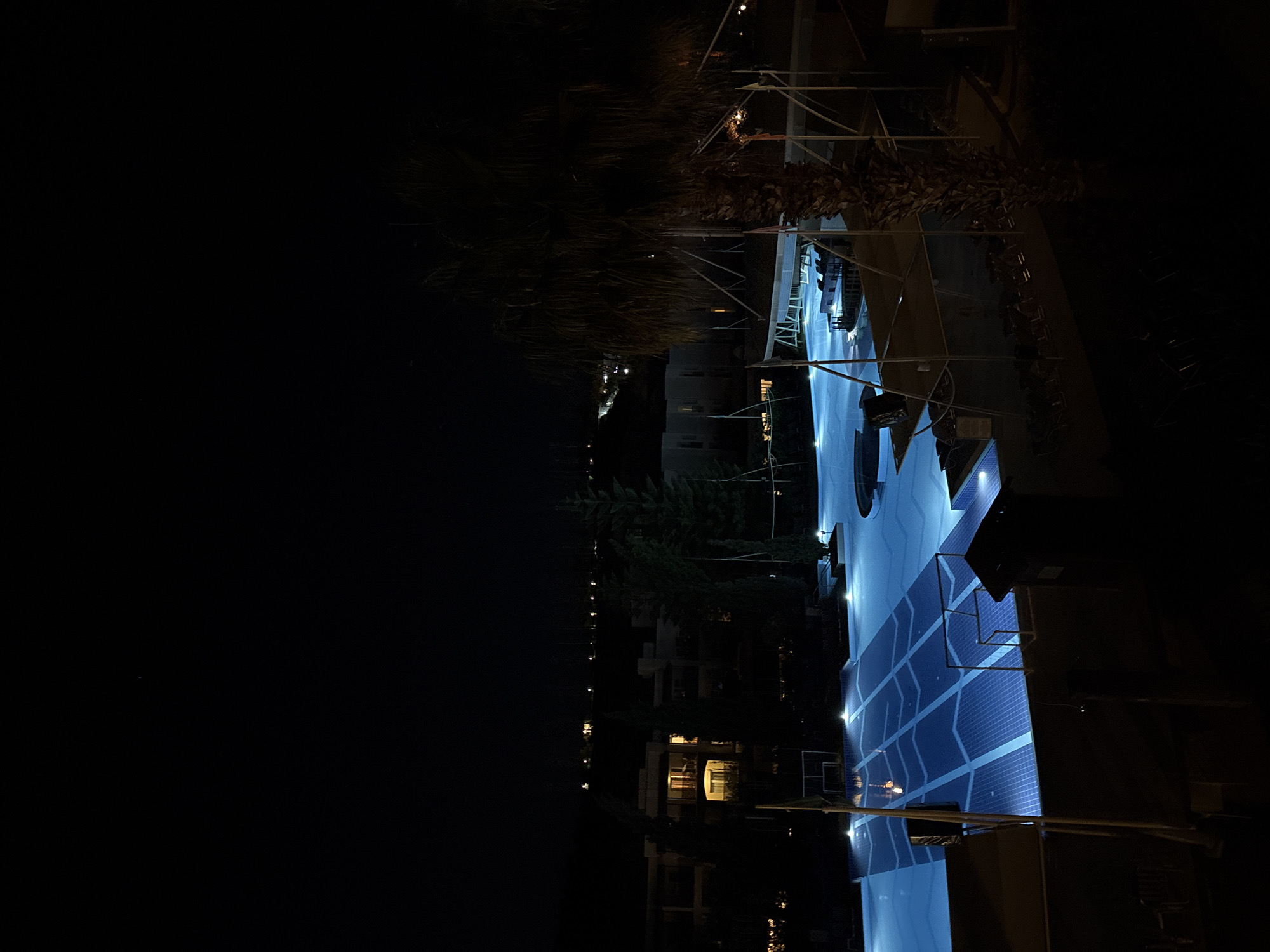 This media file captures a tranquil night scene, likely at a resort or hotel in Selçuk, Turkey. The central and most illuminated feature is a large swimming pool filled with light blue water, glowing brightly against the dark surroundings. The bottom of the pool displays a distinct geometric tiled pattern, formed by darker blue shapes resembling chevrons or arrows, creating visual interest. Steps or ladders are visible on the right side of the pool, providing access.
Around the pool, various dark structures, possibly part of a deck, railings, or architectural elements, are faintly outlined by the pool's light. To the left of the pool, tall palm trees and other dark foliage are silhouetted against the dark sky, contributing to a tropical or Mediterranean atmosphere. In the background, several multi-story buildings can be discerned, with a few windows emanating a warm yellow glow, suggesting occupied rooms or interior lighting. Further in the distance, a scattering of small, bright lights forms a horizontal line, indicating the presence of a town or city extending into the dark horizon.
The image was taken at night, characterized by the deep black sky and the reliance on artificial lighting from the pool and buildings. The weather appears clear. No people are visible, implying a quiet, late-night moment at the location. There is no visible text present in the image. The overall scene conveys a sense of peacefulness and calm.