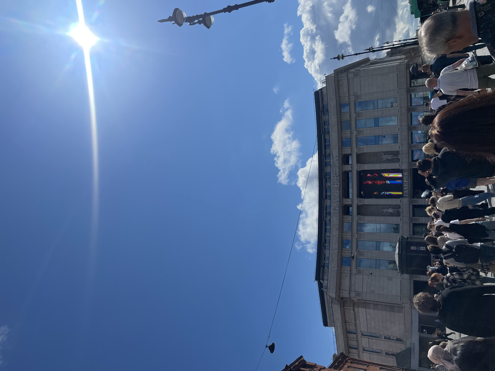 The image depicts the Nevsky Centre shopping mall, also known as Galeria, located on Nevsky Prospekt in Saint Petersburg, Russia, under a clear blue sky with scattered white clouds. A bright sun flares in the upper right corner of the frame, creating a lens flare effect. The facade of the Galeria building is visible, featuring classical architectural elements, large windows, and signage. In front of the building, a crowd of people is gathered, suggesting a busy urban environment. Various individuals are seen walking, standing, and interacting in the foreground and midground. The scene captures a typical daytime view of a popular public space in the city.