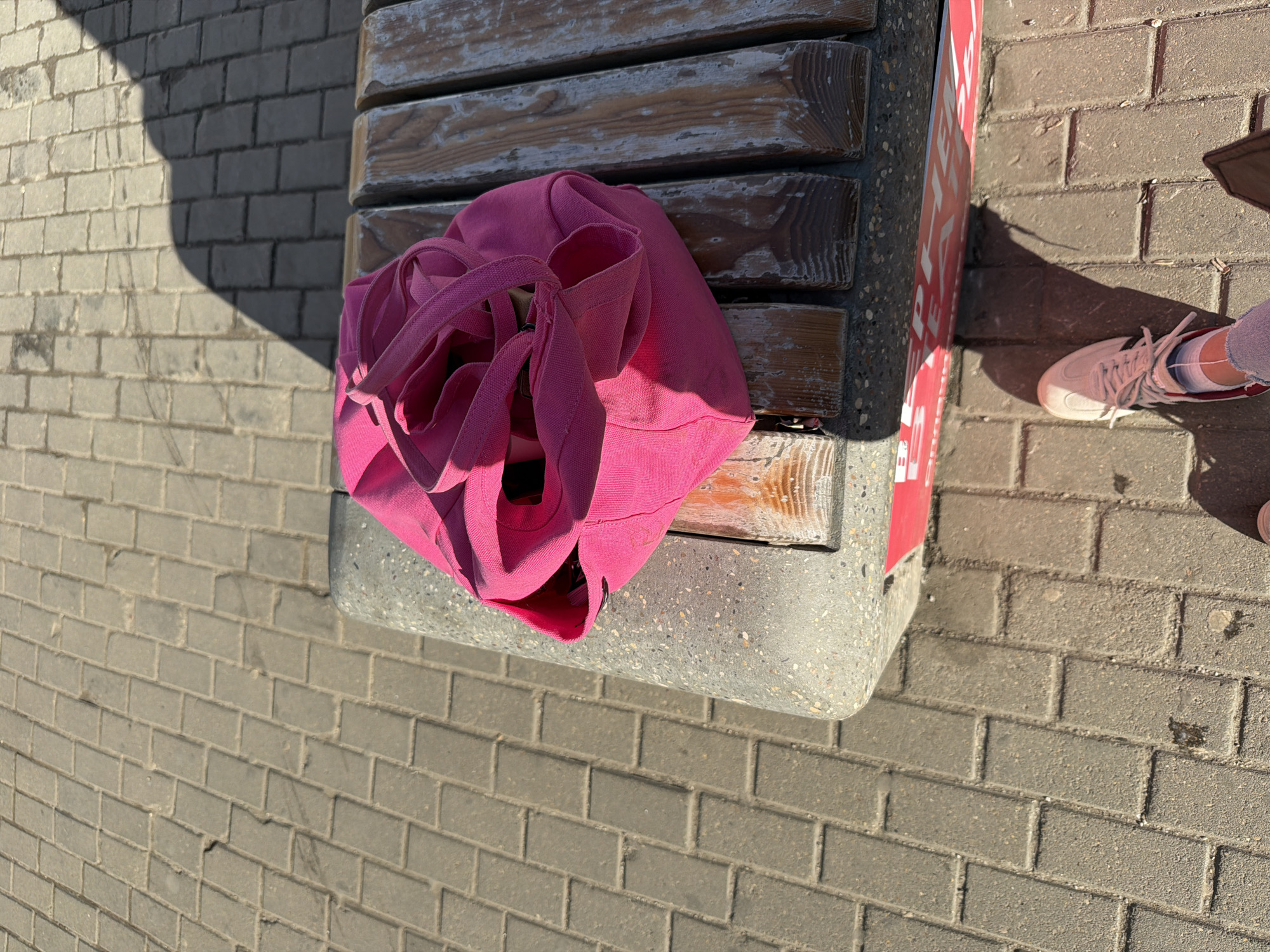 The scene is outdoors, on a paved surface with brick-like stones, likely a sidewalk in a city. A wooden bench with a concrete base and a red/white vertical panel is visible. A bright pink tote bag is resting on the bench. Part of a person's leg, wearing a white sneaker with red details and white socks, is visible to the right of the bench. The image is taken with a slight downward angle. Given the context clues, and the style of benches often found in Moscow, Russia, the location is likely somewhere in that city.