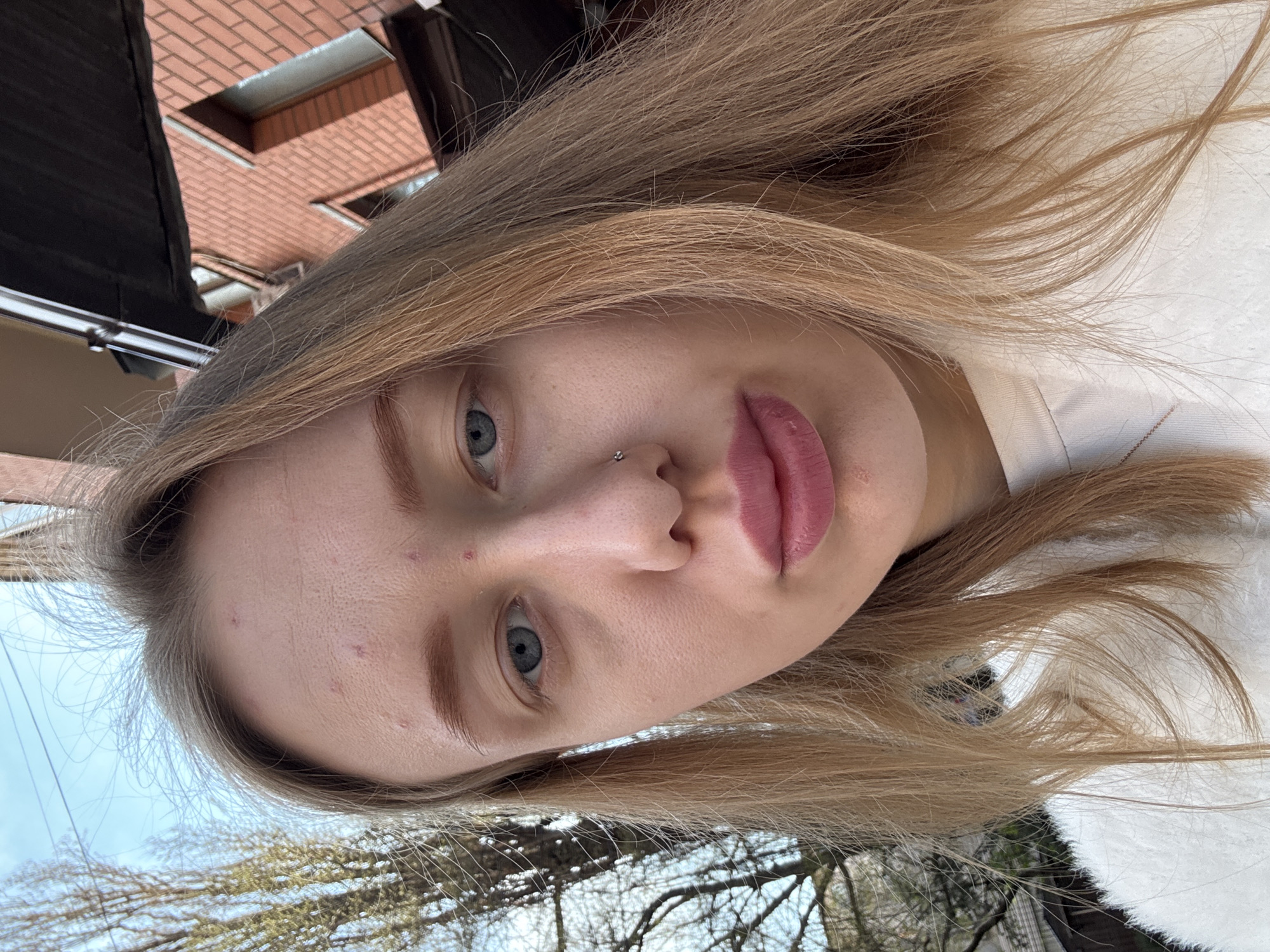 A medium close-up photograph depicts a blonde woman with blue eyes, a nose ring, and light pink lips. She is looking directly at the camera. Her complexion is fair, with a few blemishes visible on her forehead. Her hair is long, straight, and a light golden color.
The background includes elements of the outdoors such as a building facade with brick work, trees with bare branches against a light blue sky. The building facade suggests an urban location. This is likely located in Rostov-on-Don, Russia.