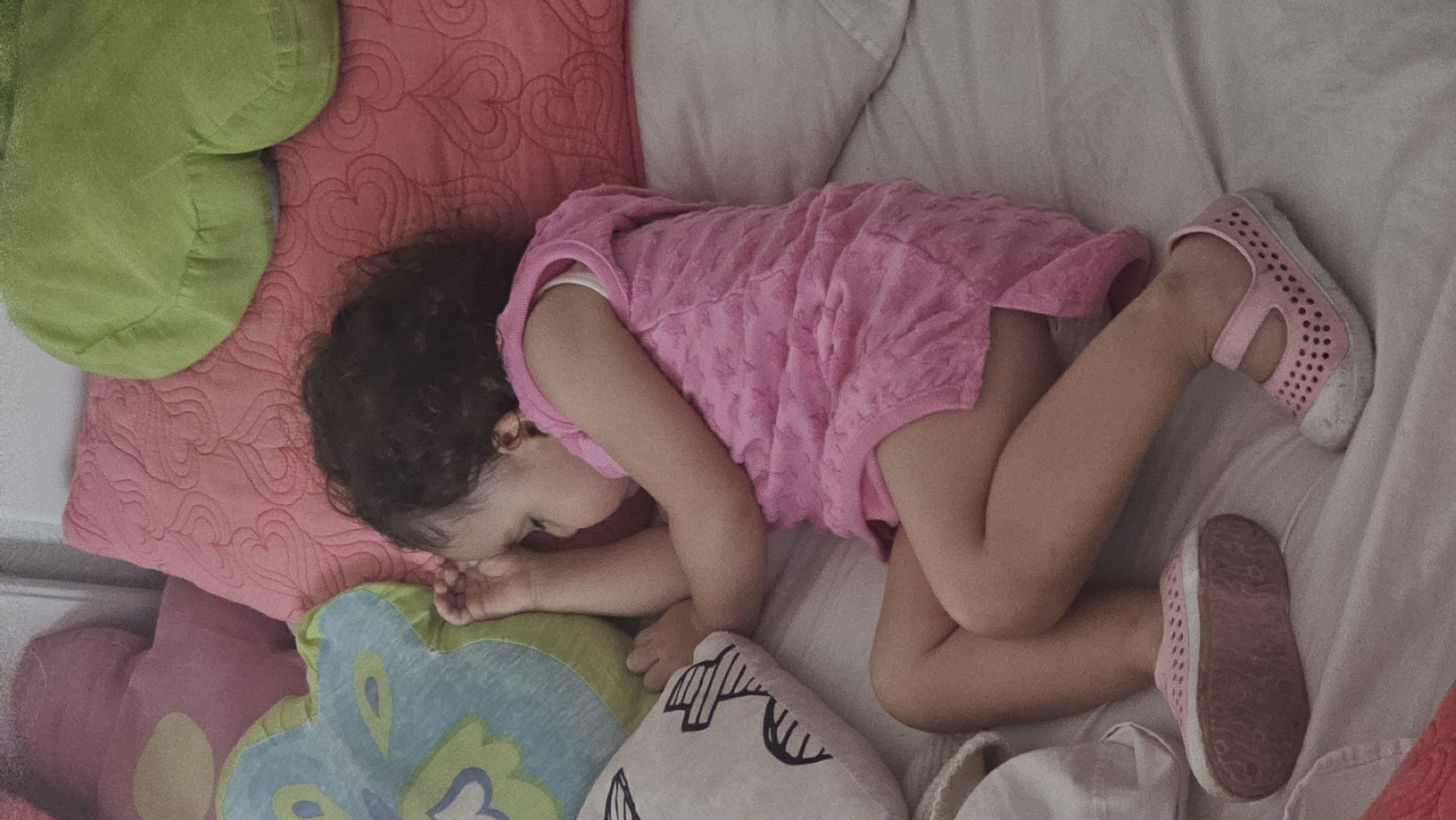 A child is lying on a bed, asleep. The child is wearing a pink dress and pink shoes, one on each foot. The bed is covered with several pillows of different colors and shapes including green, pink with heart patterns, and blue with a floral design. The bedding appears to be white. The location is presumed to be in a private residence in San Bernardino Tlaxcalancingo, Mexico.