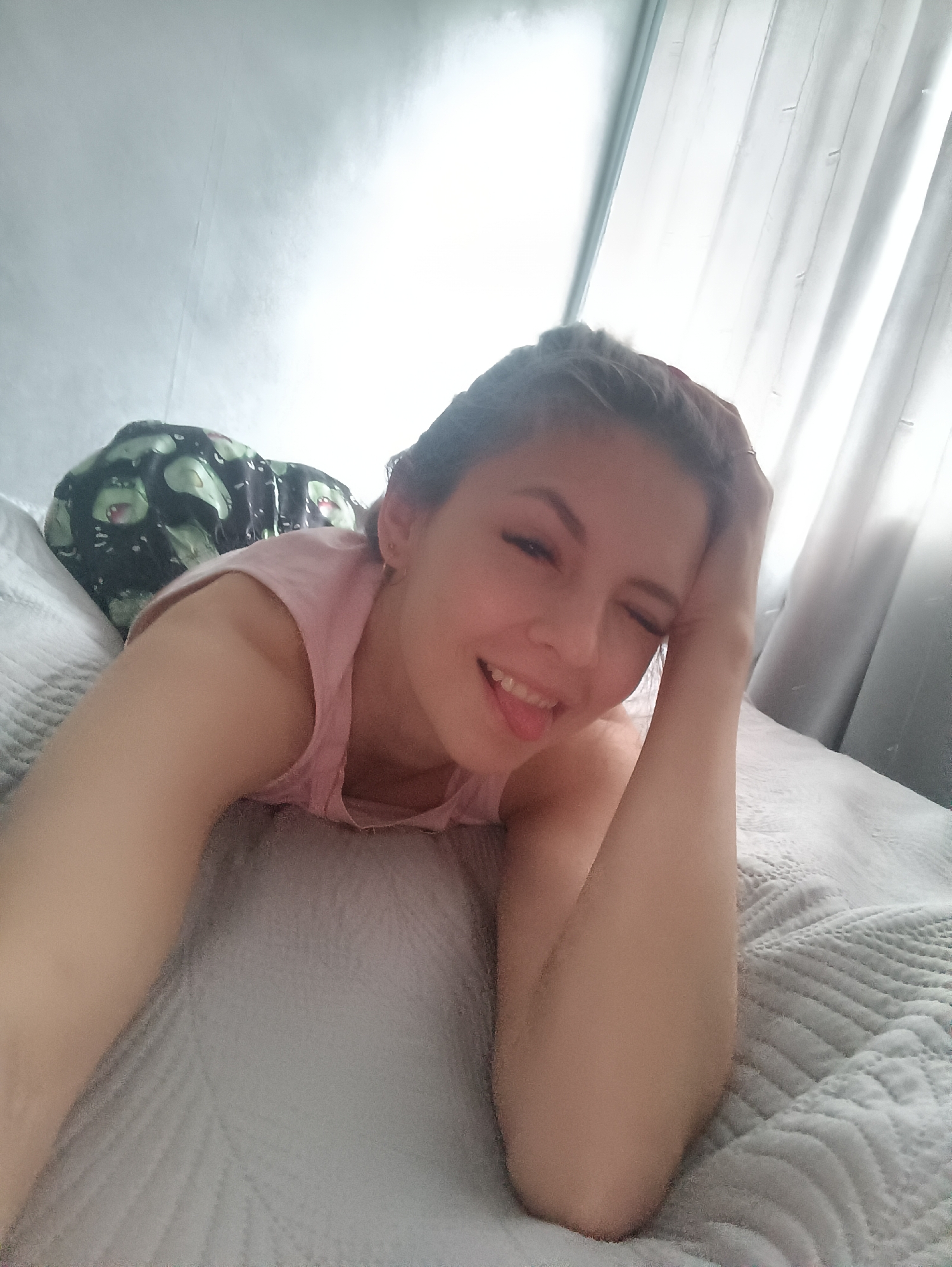 A young woman lies on a white, textured bed with her head propped up on her hand. She is wearing a pink tank top and patterned pajama pants with avocado designs. Her tongue is slightly out, and she is winking. Behind her is a white wall and a window covered by gray curtains. The location is Unknown, Unknown.