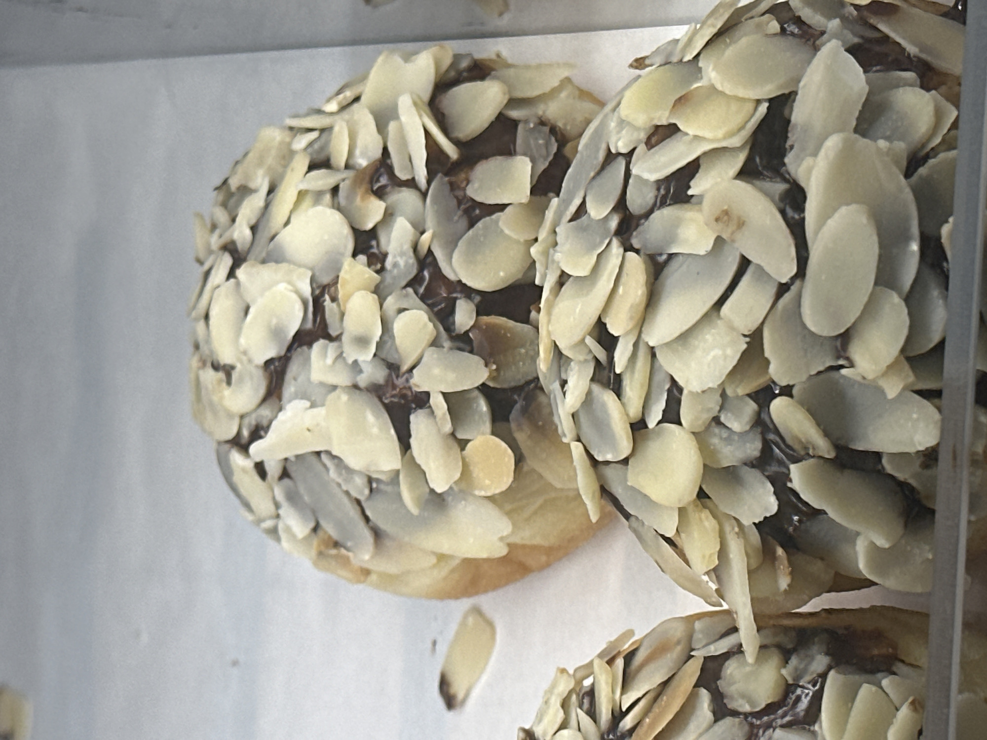 The image displays a close-up, slightly overhead view of two almond-covered pastries, likely a type of sweet bun or cake. Each pastry is densely topped with light-colored, slivered almonds, which cover a dark, possibly chocolate or nut-based, filling or glaze. The underlying pastry base, visible at the edges of the almond coating, appears golden brown and slightly flaky.
The pastries are arranged on a white surface, possibly baking paper or a tray, with a plain, light grey background suggesting they are displayed in a bakery, cafe, or dessert shop. The lighting is bright, highlighting the texture and individual flakes of the almonds. A third pastry is partially visible in the bottom right corner, indicating multiple items are present. The overall impression is one of an appealing food display. There is no visible text or specific location cues within the image, though the provided context suggests the location is Fethiye, Turkey. No people or specific activities are depicted beyond the presentation of the food.