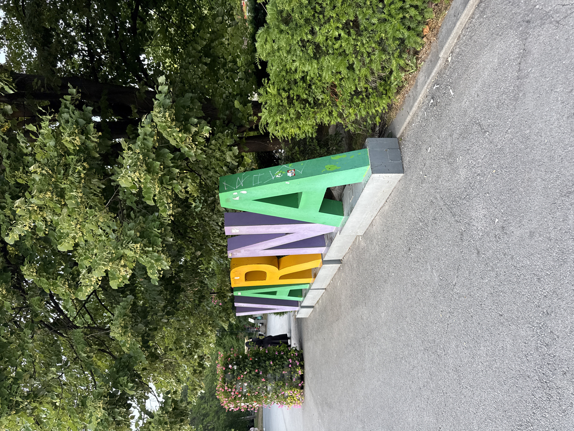 The image depicts the colorful letters spelling "VARNA" in large, three-dimensional block letters, positioned outdoors on a paved walkway. The letters are arranged sequentially from left to right: V (purple), A (green), R (yellow), N (purple), A (green). The letters are resting on concrete bases.
To the left of the "VARNA" sign, there is a tall, cylindrical planter overflowing with pink and white flowers. Behind the planter, a person dressed in a long black garment, possibly a dress or robe, is walking away from the camera. Further in the background, more trees and greenery are visible.
The environment appears to be a park or public space with a paved pathway in the foreground and lush trees providing shade. The lighting suggests it is daytime, with sunlight filtering through the leaves. The scene is clean with no visible litter except for a few cigarette butts scattered along the edge of the pathway near the right side of the image. The overall impression is a pleasant, outdoor urban setting.