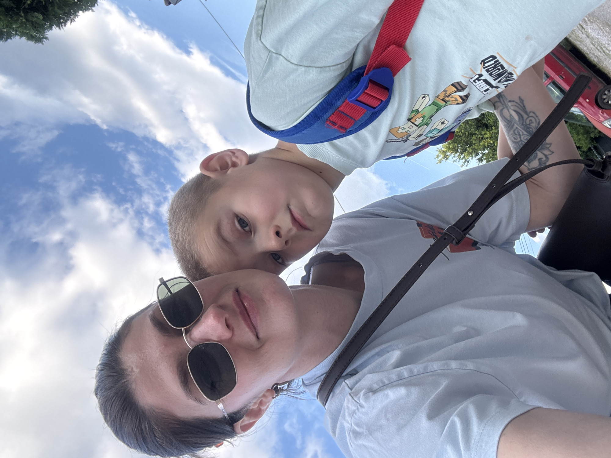 A rotated, slightly tilted, eye-level shot features a woman and a young boy in an outdoor setting, likely during the daytime.
The woman, positioned at the bottom, wears sunglasses and a light blue t-shirt with a dark brown strap across her chest. Her dark hair is pulled back.
The young boy is above her, wearing a light-colored t-shirt with a Minecraft character design and a red and blue backpack.
The background shows a partly cloudy sky and glimpses of green trees, as well as the top of a red car.
Given the architecture, street layout, vegetation, and vehicle, it is not possible to determine if this scene occurred in Podgorica, Montenegro, without further information.