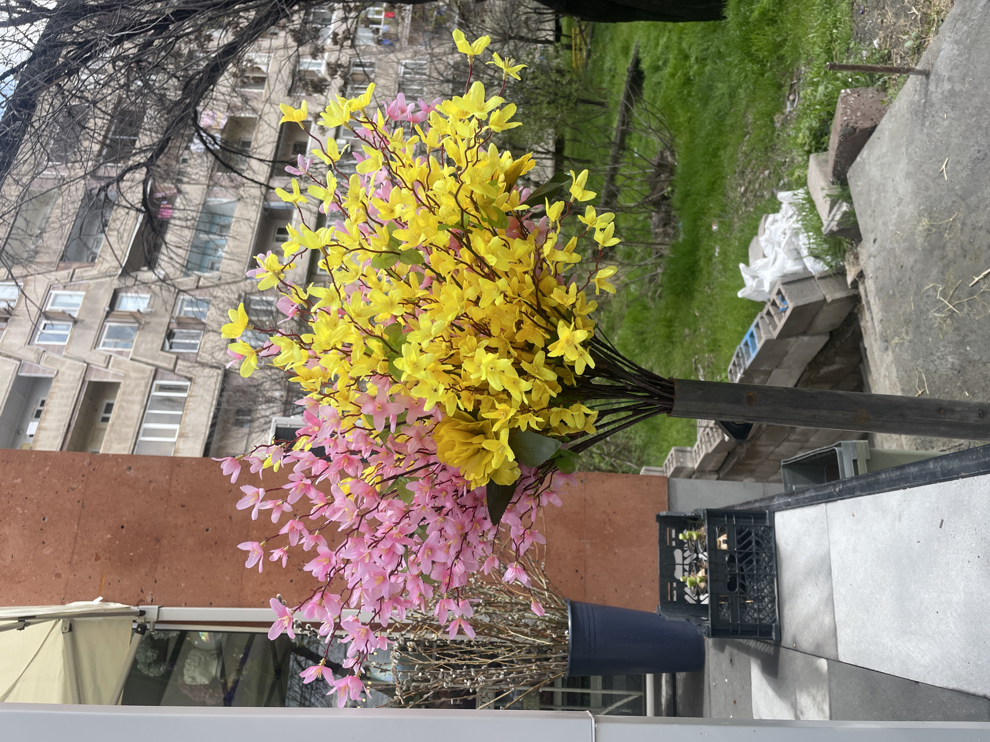The image depicts a flower arrangement in Yerevan, Armenia. The arrangement features yellow and pink blossoms in a container. Behind the flowers is a cityscape with a tall building visible. In the background is a patch of green grass. There are stone blocks and debris on the right side of the picture. There is a black crate with some plants in it.