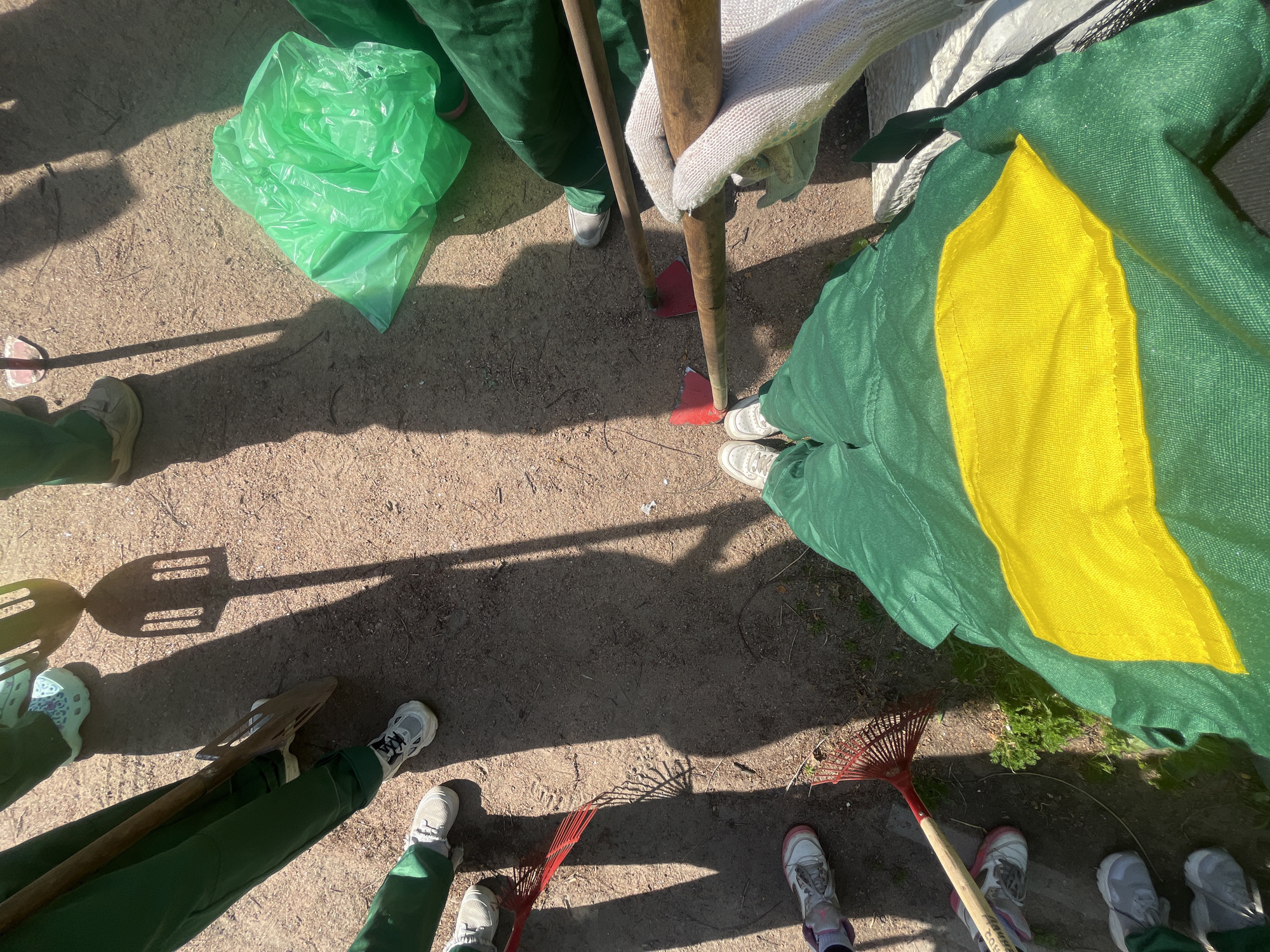 This image, taken from a first-person perspective, depicts a group of individuals dressed in green work uniforms, gathered on a dirt and gravel surface. The setting appears to be outdoors, possibly in an urban park or construction site in Saint Petersburg, Russia, suggested by the presence of several tools and gardening equipment.
In the foreground, the legs and feet of multiple individuals are visible, clad in green pants and athletic shoes. One person in the center wears a green garment with a prominent yellow stripe across the lower half. Various tools are scattered around the scene. To the left, two red rakes lie on the ground. Further up, a shovel with a wooden handle is held by an unseen person. In the background, a bright green plastic bag is visible, along with other shovels and tools casting long shadows on the ground due to the bright sunlight. The overall impression is that of a group engaged in outdoor work or preparing for such an activity.