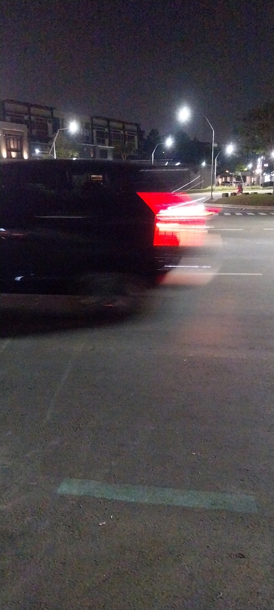 This is a low-light, long-exposure photograph taken at night on a street in the city of Medan, Indonesia. The primary subject is the taillight of a dark-colored vehicle, captured as a streak of red light due to motion blur. The vehicle appears to be moving from left to right across the frame. In the background, modern multi-story buildings with prominent architectural features are visible, illuminated by streetlights. The environment includes a paved road with visible lane markings and a pedestrian crossing. Streetlights are present along the road, creating pools of light. The overall scene suggests an urban environment at night, with the motion blur indicating activity and traffic.