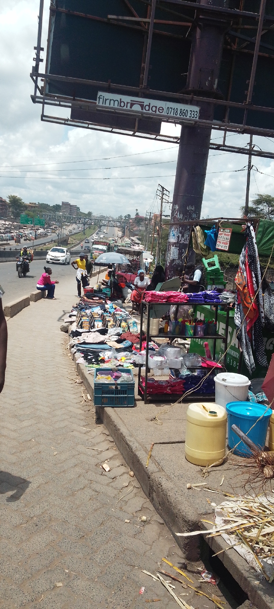 The image shows a bustling roadside scene in Nairobi, Kenya, with a congested highway visible in the background. A billboard with the word "firmbridge" and a phone number "0718 860 333" stands above a makeshift market along the side of the road. Several vendors and customers are present, engaging in trade. Merchandise including clothing, household items, and other goods is displayed. The street is cluttered, with containers and other debris littering the ground near the sidewalk.
