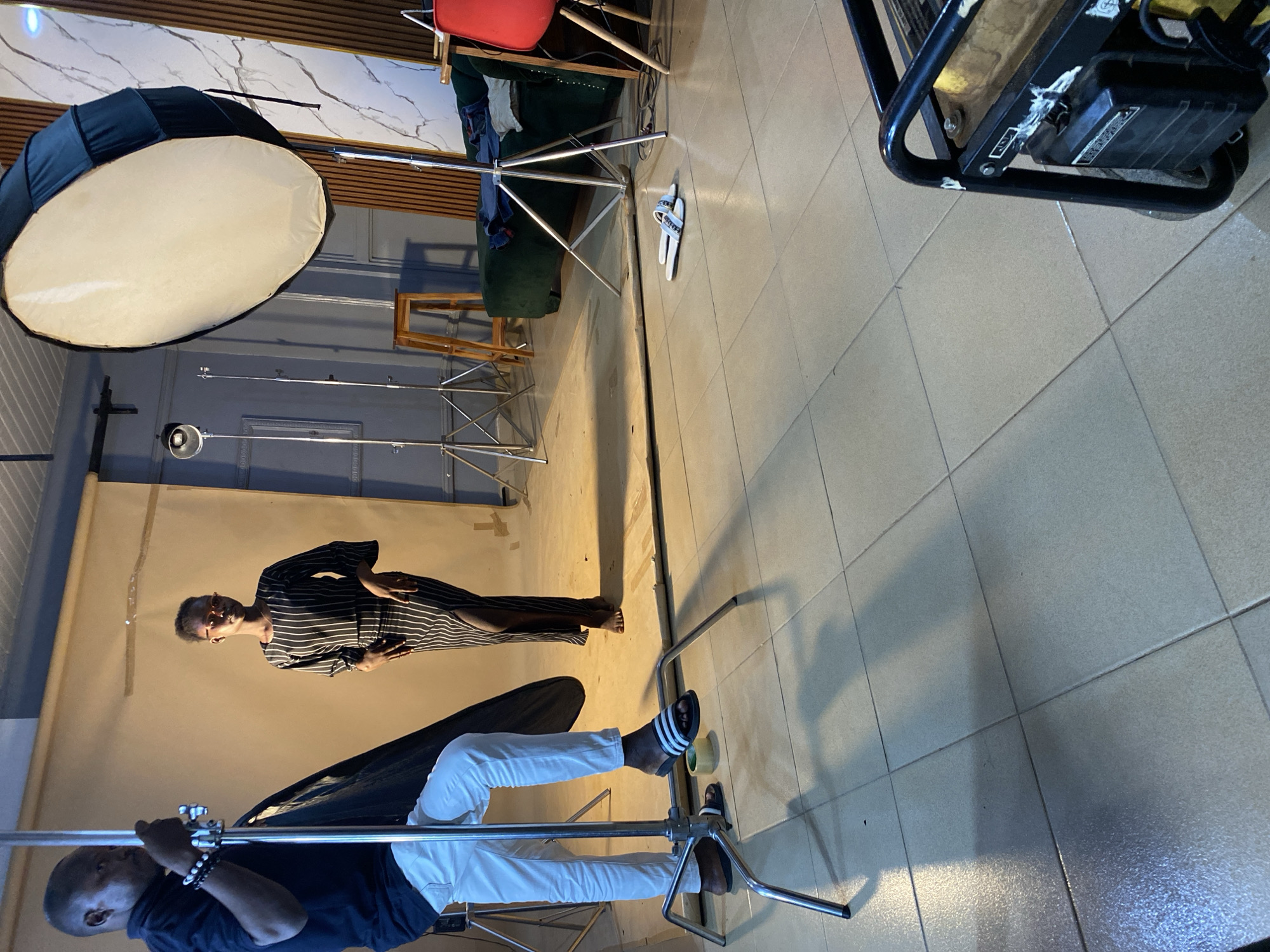 The image shows a photo shoot in what appears to be a studio setting. A woman in a black and white striped dress poses in front of a cream-colored backdrop. Two individuals are likely part of the crew; one is visible adjusting equipment, dressed in a black shirt and white pants. Various lighting equipment, including a large softbox and light stands, are present. The floor is tiled. Given the prompt context, the location is presumed to be in Bukuru, Nigeria.