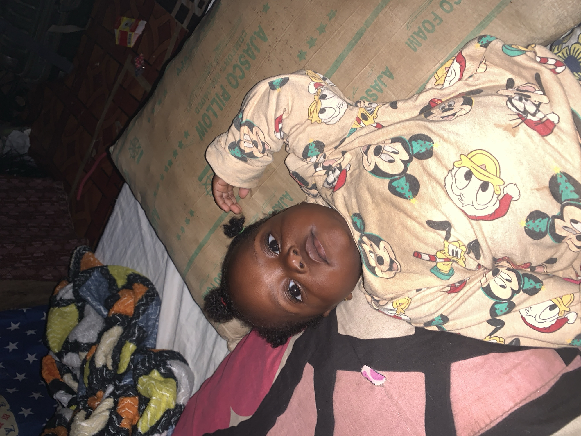 The image captures a close-up, overhead shot of a dark-skinned baby girl with two small pigtails, lying on a bed in Agban, Nigeria. She is wearing a beige pajama set adorned with cartoon characters, including Mickey Mouse and Donald Duck. The baby is positioned on a large, brown cardboard pillow that appears to be part of a mattress packaging, as indicated by the text "AJASCO PILLOW" and "CO FOAM" visible on its surface. To the left of the baby, a pink and black patterned pillow is partially visible. In the background, a colorful, abstractly patterned blanket is draped over the bed, and beyond that, a dark, tiled floor can be seen, along with some indistinct objects, possibly part of a room's furnishings. The lighting suggests an indoor setting, likely at night or in a dimly lit room.