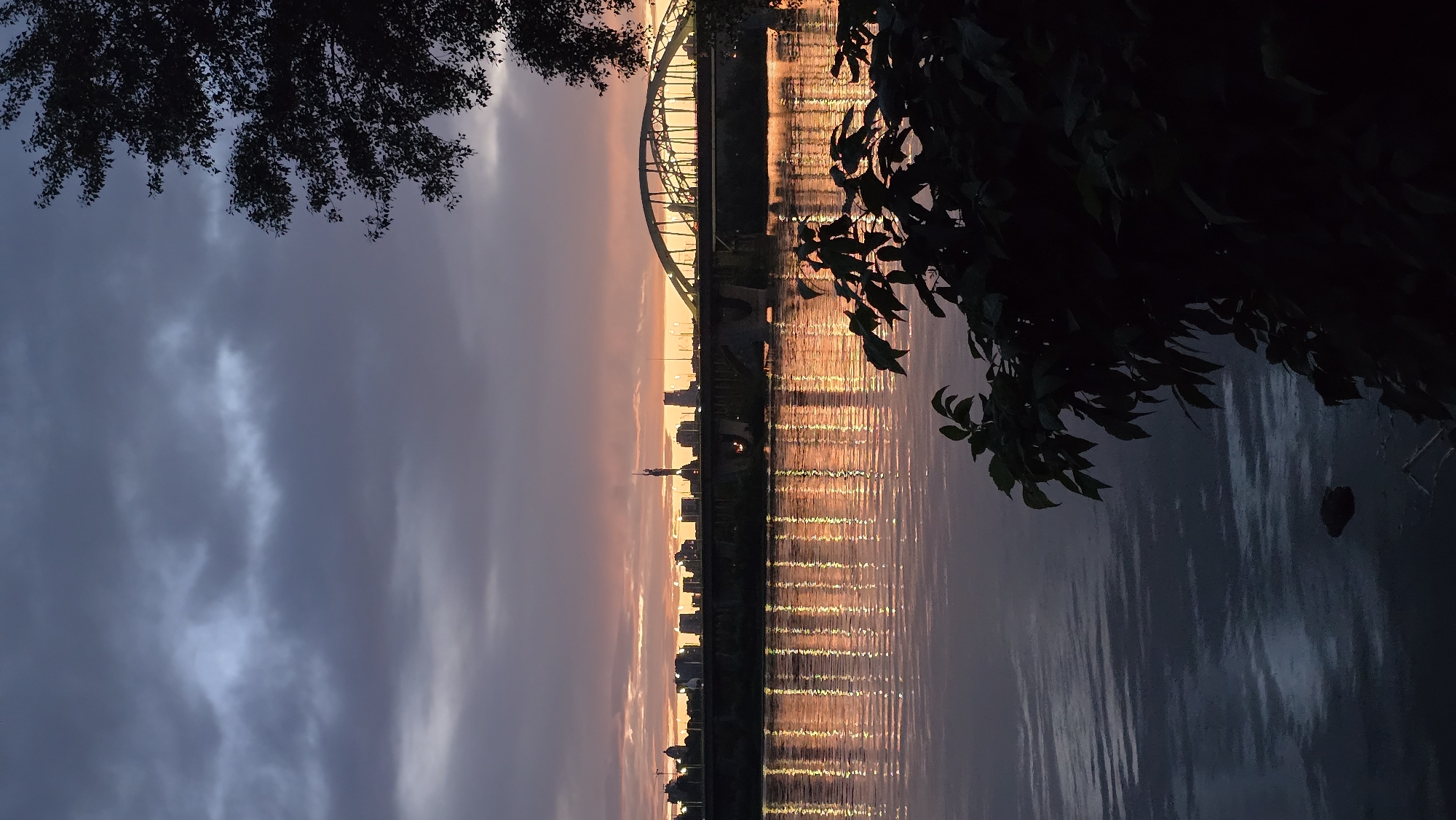 The image depicts a silhouetted cityscape of Kyiv, Ukraine, as seen across a body of water during a twilight sunset. The sky is filled with dramatic clouds, displaying hues of purple, gray, and pink from the setting sun. A prominent arched bridge spans the river, with its silhouette clearly defined against the colorful sky. The cityscape consists of various buildings and structures, their forms obscured by the low light, with a tall monument or spire visible in the center of the skyline. The water in the foreground reflects the vibrant colors of the sky and the lights from the city and bridge, creating a shimmering effect. On the right side of the frame, the dark silhouette of foliage from a tree is visible, framing the scene and adding depth. The overall atmosphere is serene and picturesque, capturing the beauty of the city at dusk.