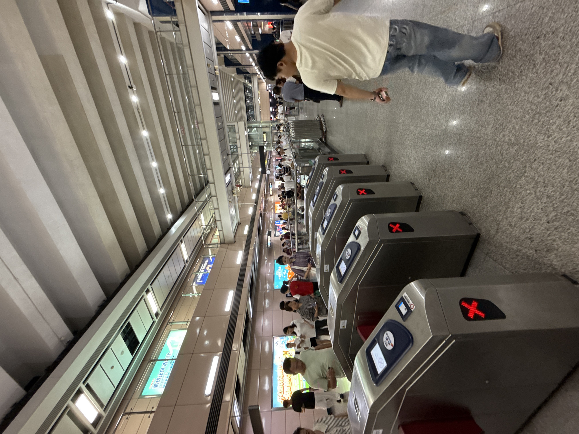 In Haizhu District, China, a crowd of people is seen passing through a metro station's ticket gates. The image focuses on the ticket gates themselves, which are silver and equipped with red "X" indicators, suggesting some are not operational or access is denied. A man in a white t-shirt and blue jeans walks away from the camera, while others are seen in the background, either queuing or moving through the station. The environment appears to be an indoor, well-lit space with a high ceiling and advertisements displayed on the walls.