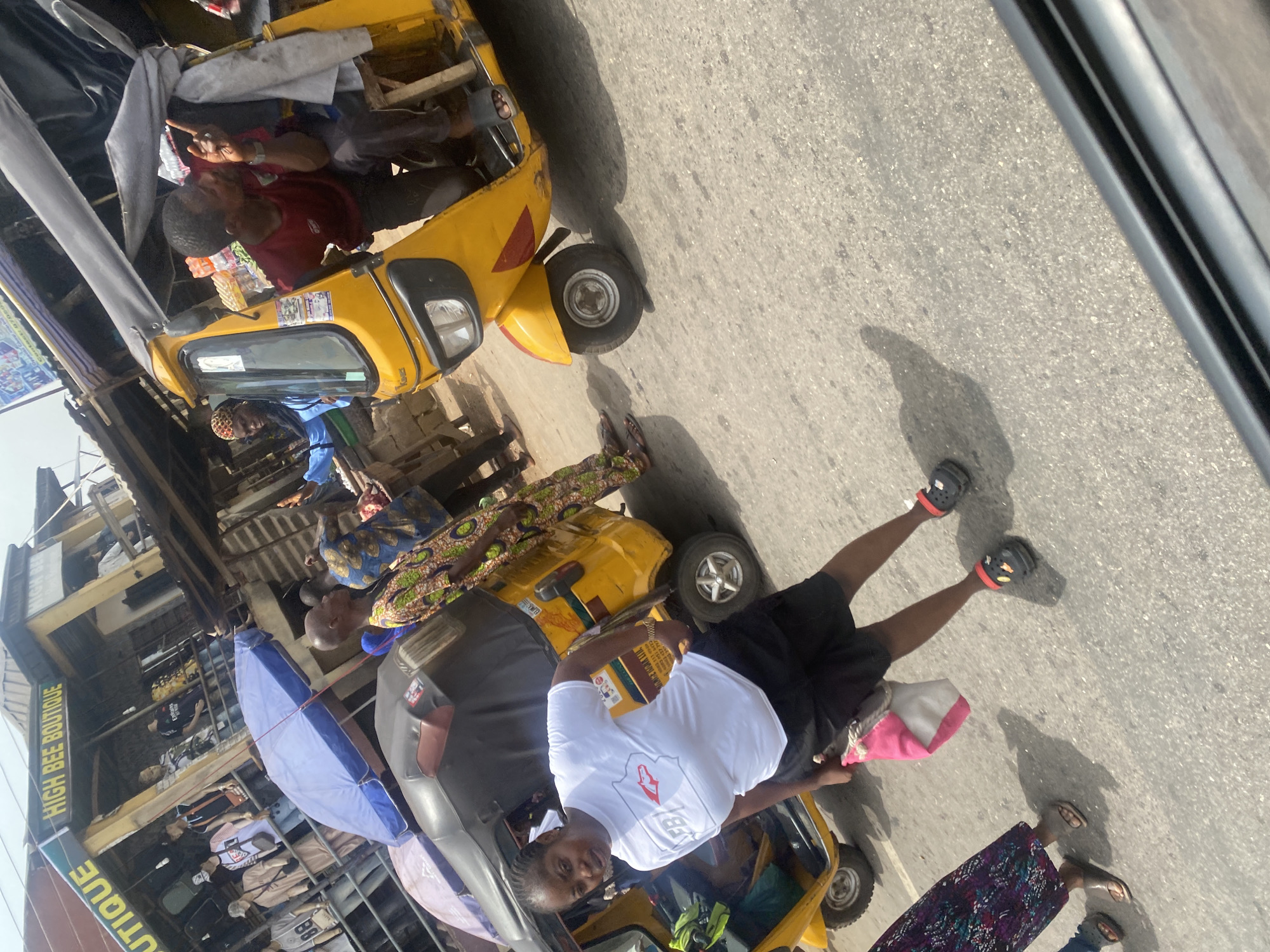 The scene shows a street in Alakija, Nigeria, with yellow auto rickshaws (tuk-tuks) being used for transportation. Several people are present; some are passengers riding in the tuk-tuks, while others are standing nearby on the street. A multi-story building with the sign "HIGH BEE BOUTIQUE" is visible in the background. The setting is outdoors on a sunny day.