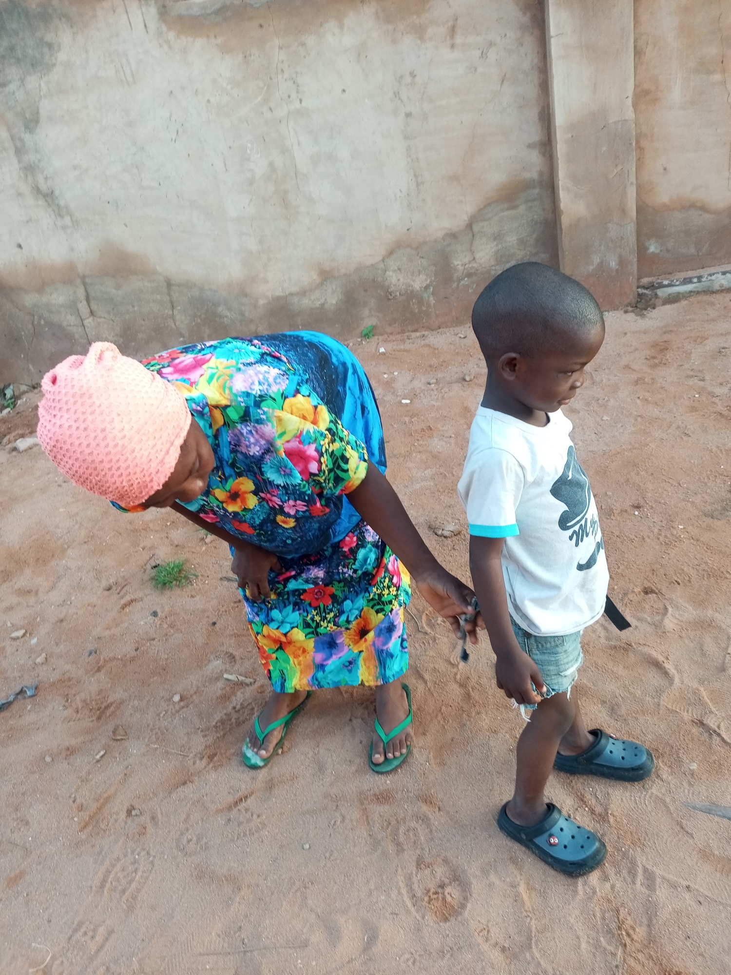 In Tempure, Nigeria, a child stands to the right of an adult in the sandy ground. The adult, wearing a pink headscarf and a vibrantly patterned dress, is bent over and appears to be holding a small object in their right hand. The child, dressed in a white t-shirt and denim shorts, looks to their right. A weathered, light-colored wall forms the background. The ground is composed of sandy dirt with scattered small stones.