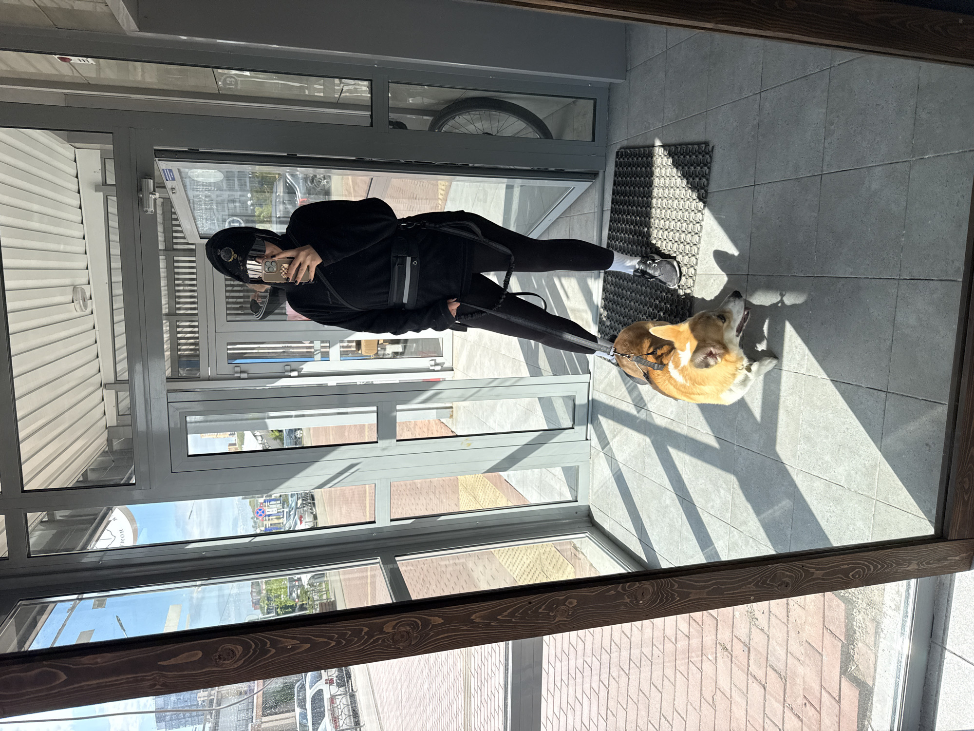 A woman in black athletic wear, a black hat, and black sneakers stands in a glass-enclosed vestibule with a tan and white corgi dog on a leash. The woman is taking a selfie with her phone. The floor is tiled. A dark brown wooden frame surrounds the glass. Outside, the cityscape is visible, including signs, buildings, and vehicles. The word "КРАСНОЯРСК" is visible on a street sign, which confirms the location as Krasnoyarsk, Russia. A bicycle wheel is visible through a window.