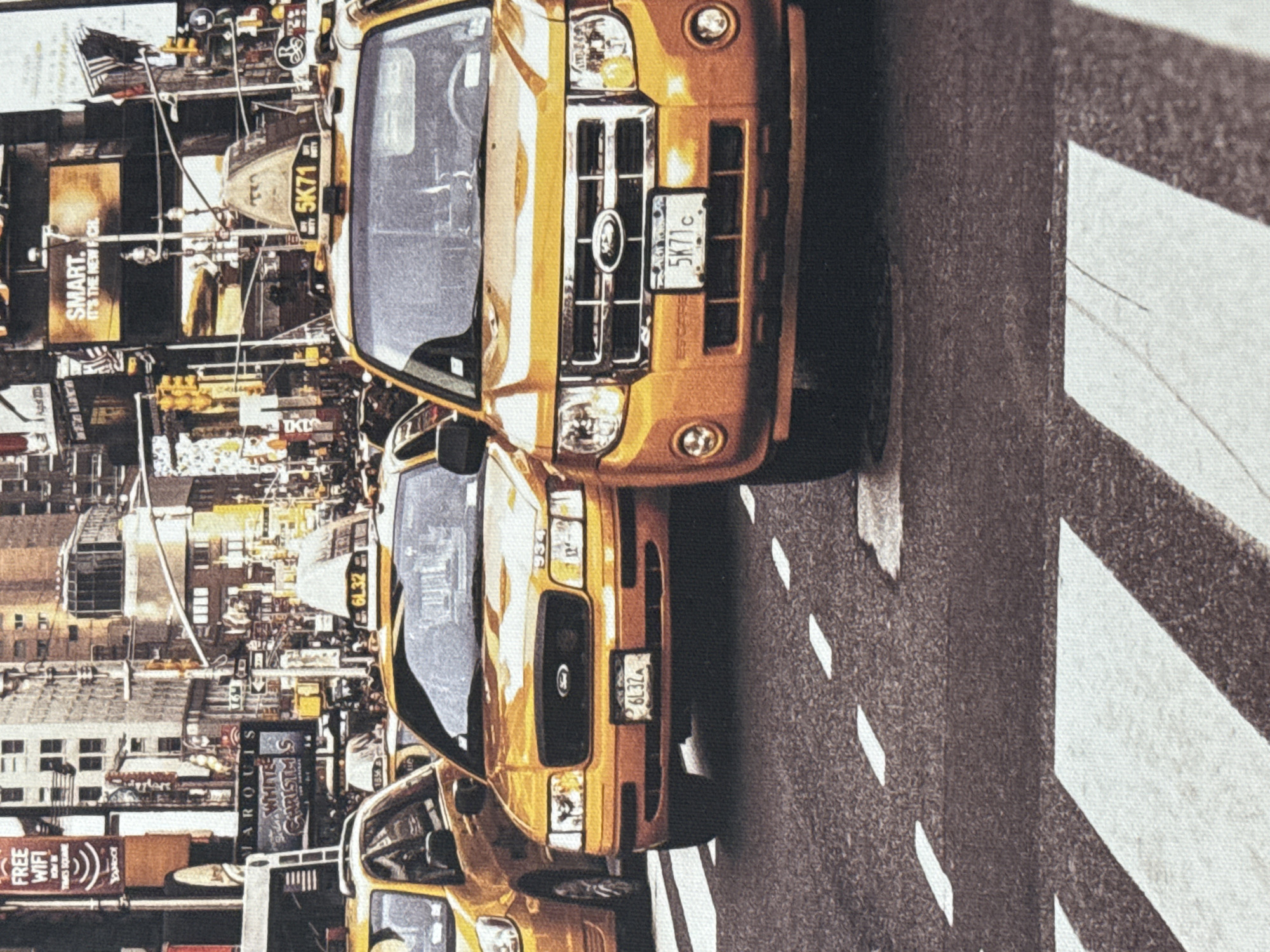 The image depicts a street scene in Times Square, New York City, with multiple yellow taxis and tall buildings adorned with numerous illuminated advertisements. The foreground features a crosswalk with white stripes. In the middle ground, two yellow taxis are prominently displayed. The taxi on the right is a Ford Escape with the license plate "5K71c". The taxi on the left is a sedan with the license plate "6L32". The background shows a bustling city environment with traffic lights, other vehicles, and large digital billboards, including one that reads "SMART. IT'S THE NEW FCK.". The scene is densely populated with signs and advertisements, characteristic of a major urban center. There are no individuals clearly visible in the image. The scene is not located in Анкудиновка, Russia.