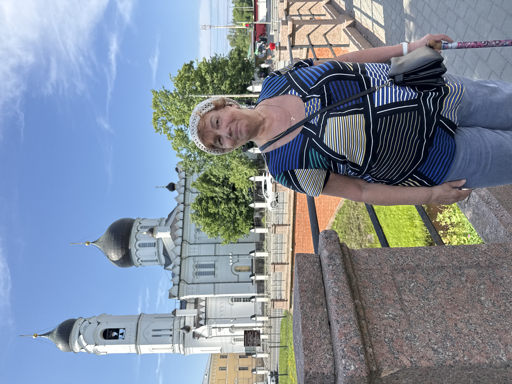 This is a vertical, eye-level photograph of a woman standing outdoors in Lomonosov, Russia. The woman, who appears to be middle-aged or older, is smiling slightly as she looks at the camera. She is wearing a white crocheted hat, a black, blue, and white striped shirt, and grey pants. She has a black purse slung over her right shoulder and is holding a cane with a patterned handle in her right hand. A gold cross necklace is visible around her neck.
Behind the woman, a large, white orthodox church with black domes and a tall bell tower is visible against a bright blue sky with wispy white clouds. Trees with green foliage are also present. The foreground features a stone structure, possibly a monument or part of the church grounds, with rough-textured pink and grey granite. The setting suggests a sunny day, likely during daylight hours. The overall mood of the image is casual and pleasant.