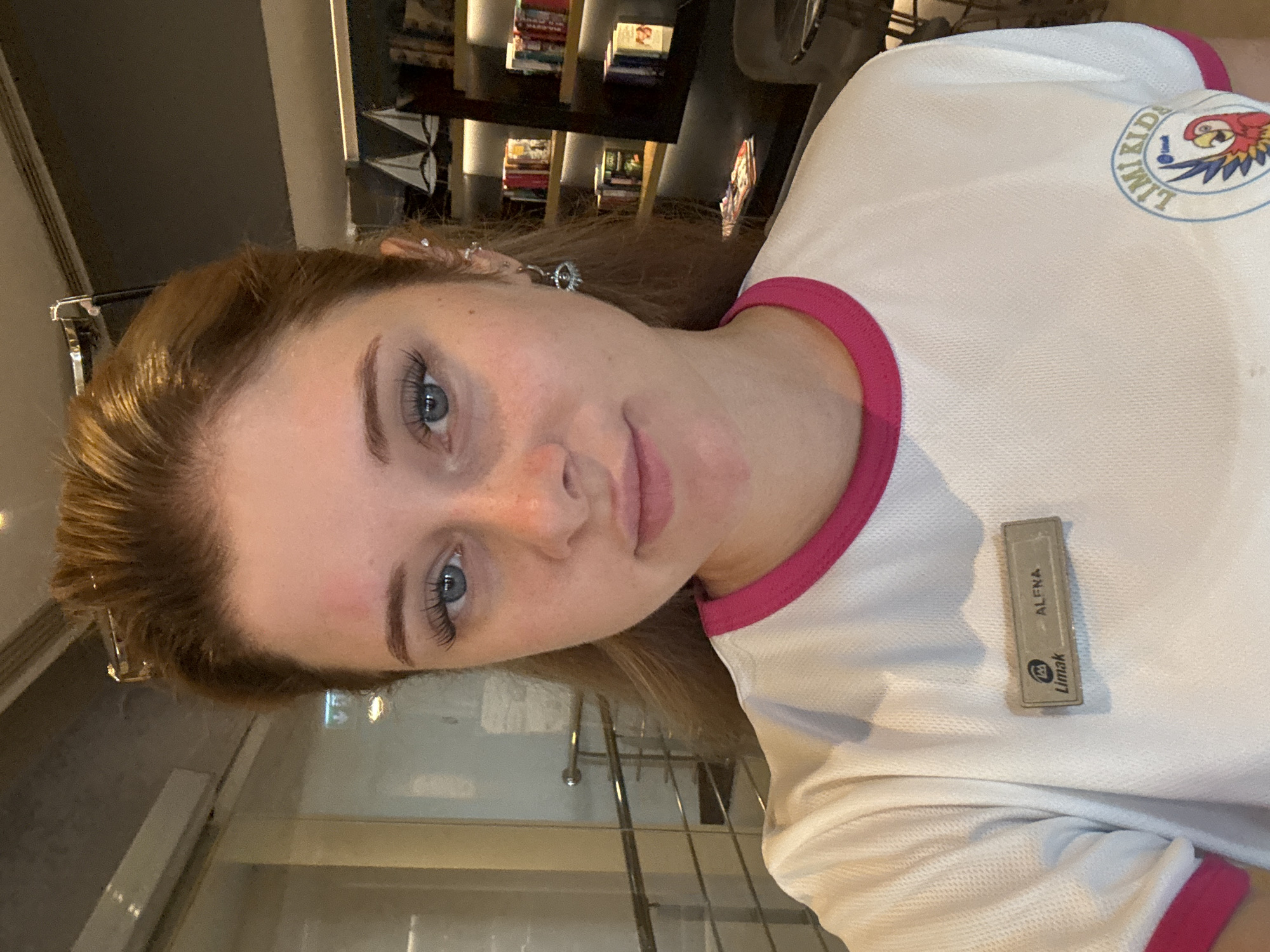 The image shows a young woman with blonde hair and blue eyes, wearing a white shirt with a pink trim and a name tag that reads "Limak ALENA". She has a red mark on her forehead above her left eyebrow and is wearing an earring in her left ear. The background appears to be a hotel lobby with bookshelves filled with books and decorative sailboats. The woman is positioned in the foreground, looking directly at the camera. Based on the "Limak" branding on her shirt and name tag, it is highly probable she is at a Limak hotel in Aksu, Turkey, a popular tourist destination known for its resorts.