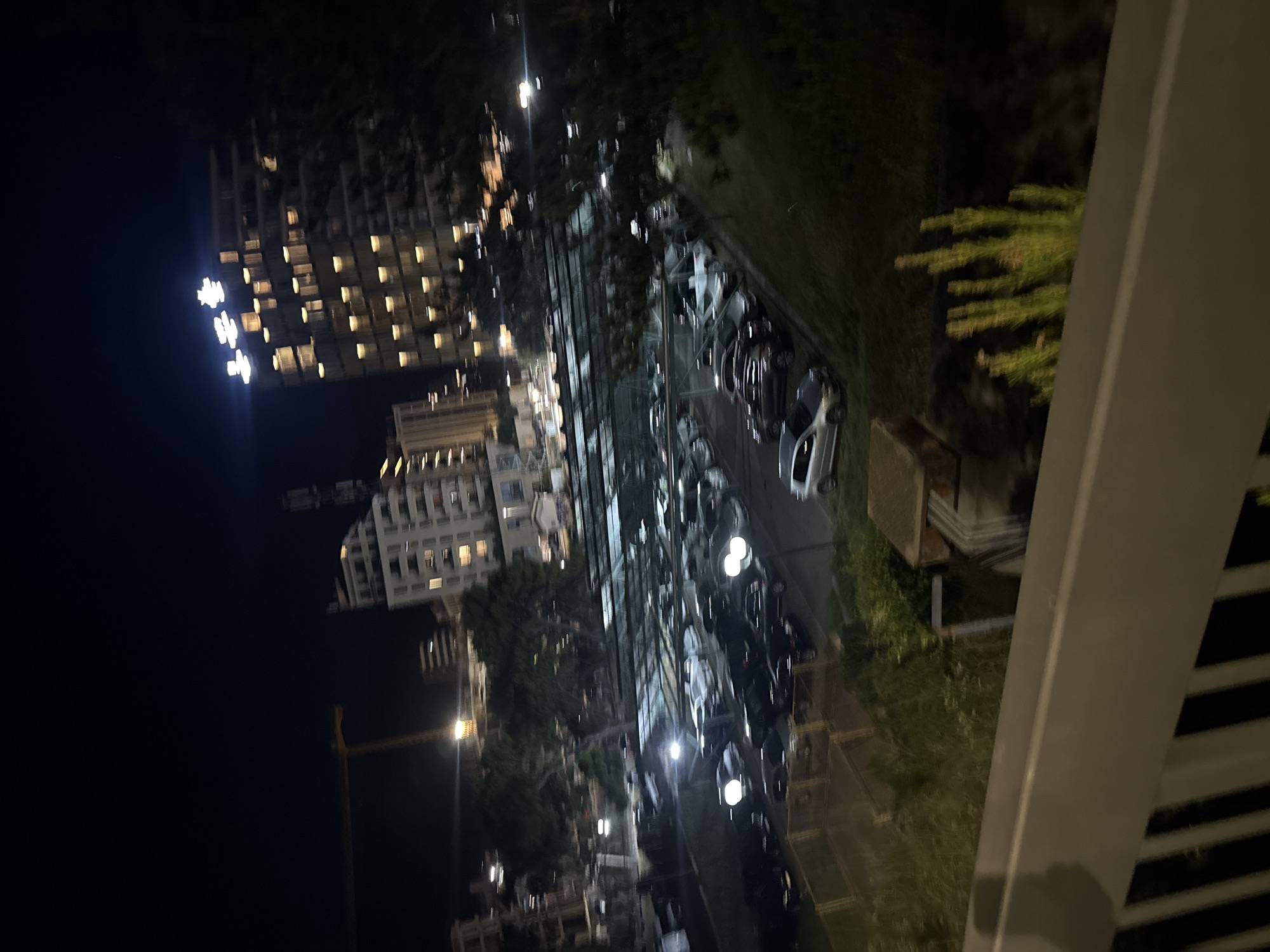 The image, taken at night in the city of Boreti, Montenegro, depicts a parking lot filled with numerous cars under artificial lighting. In the background, several modern buildings are visible, including one with illuminated signage at its peak and another with a construction crane positioned nearby. The foreground shows a portion of a railing and some vegetation. The scene appears to be a typical urban environment at night.